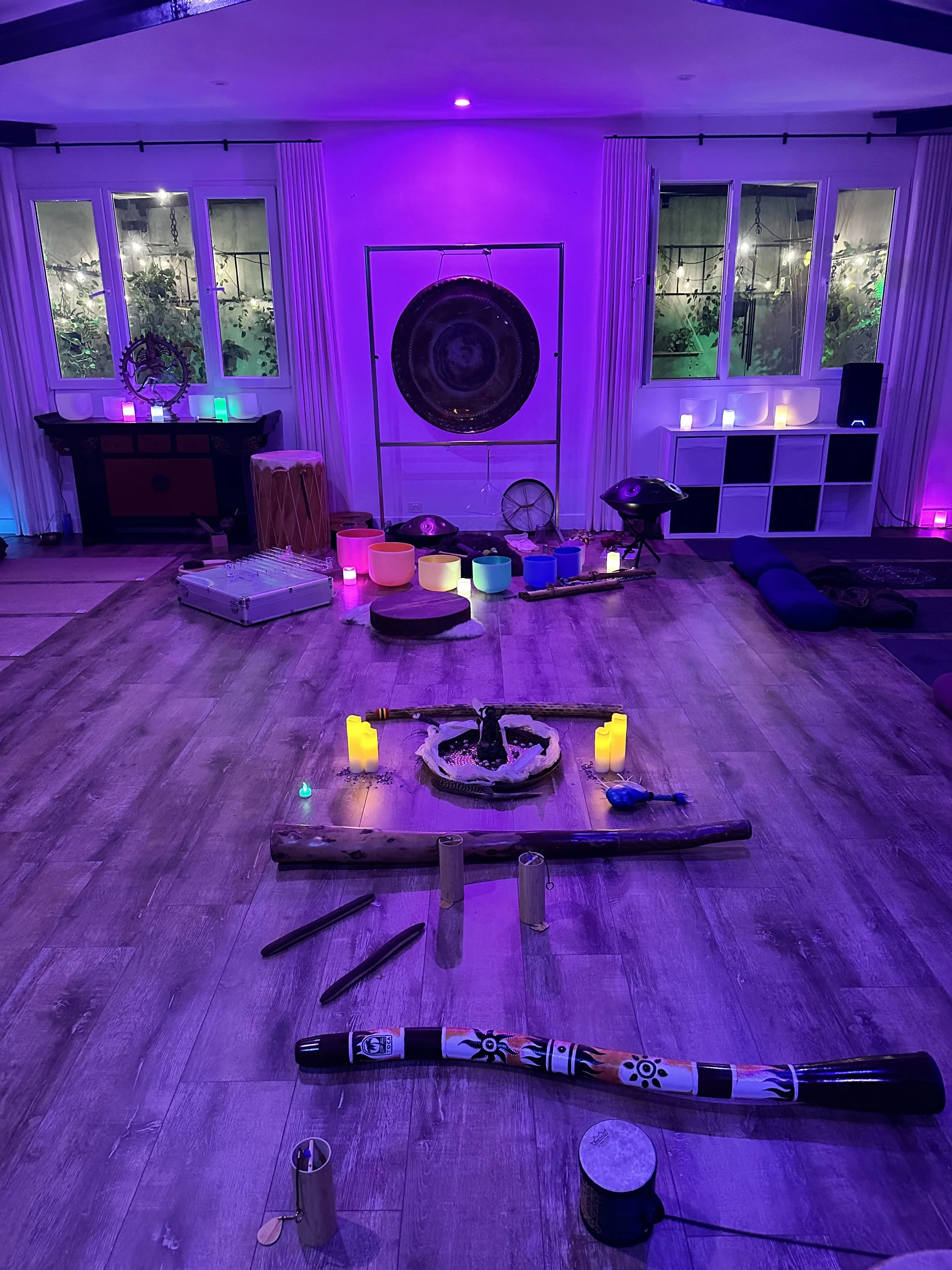 A room with purple and blue lighting, decorated with various musical instruments and meditation and sound therapy equipment, including singing bowls, chimes, drums, a gong, and candles arranged on the floor, indicating a space for sound healing or meditation.