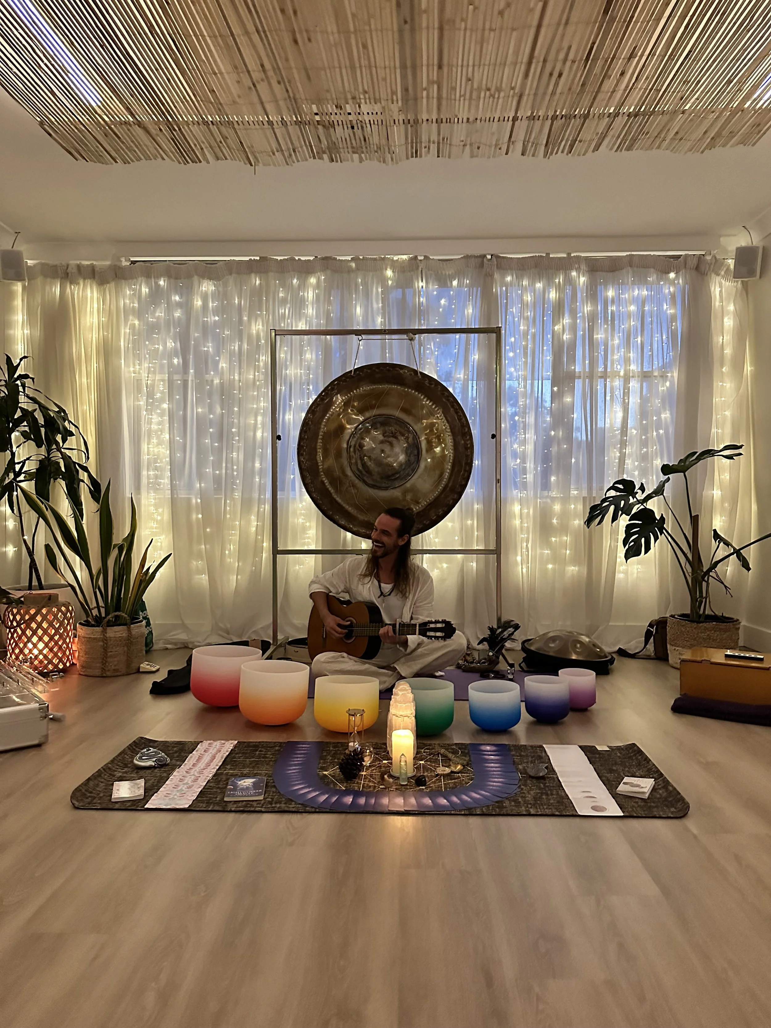 A person sitting cross-legged on a yoga mat, playing a guitar in a cozy, candlelit room decorated with plants, rainbow-colored crystal bowls, and fairy lights, with a large gong hanging on the back wall.
