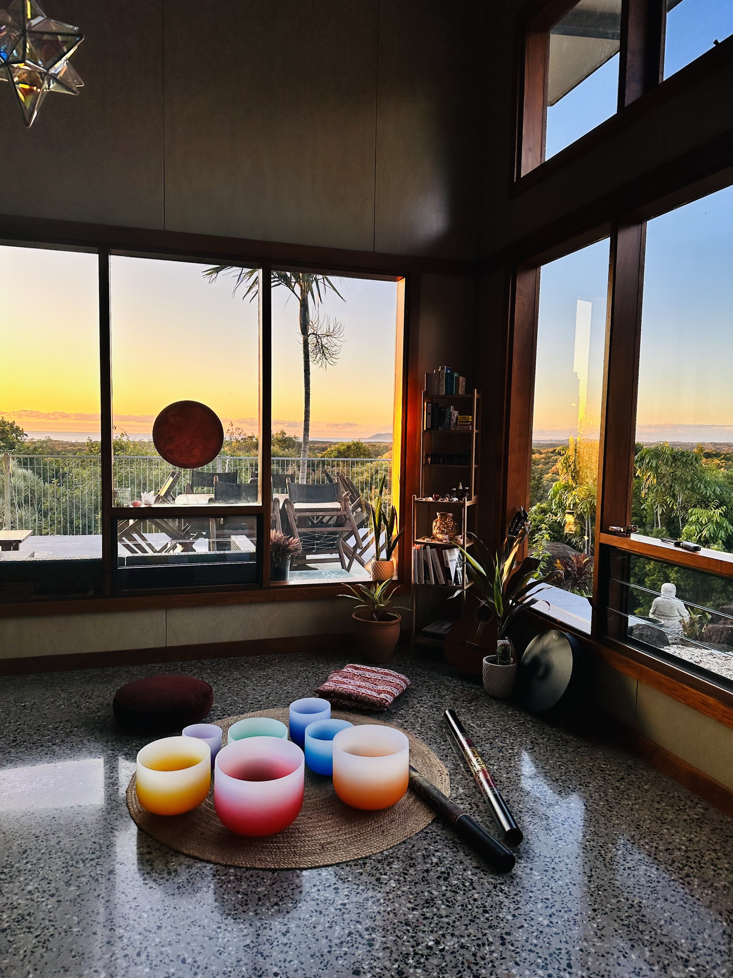 Sunset view through large windows overlooking greenery and a balcony with chairs, seen from inside a cozy room with a round mat, singing bowls, and incense on a speckled countertop, with books and plants inside.