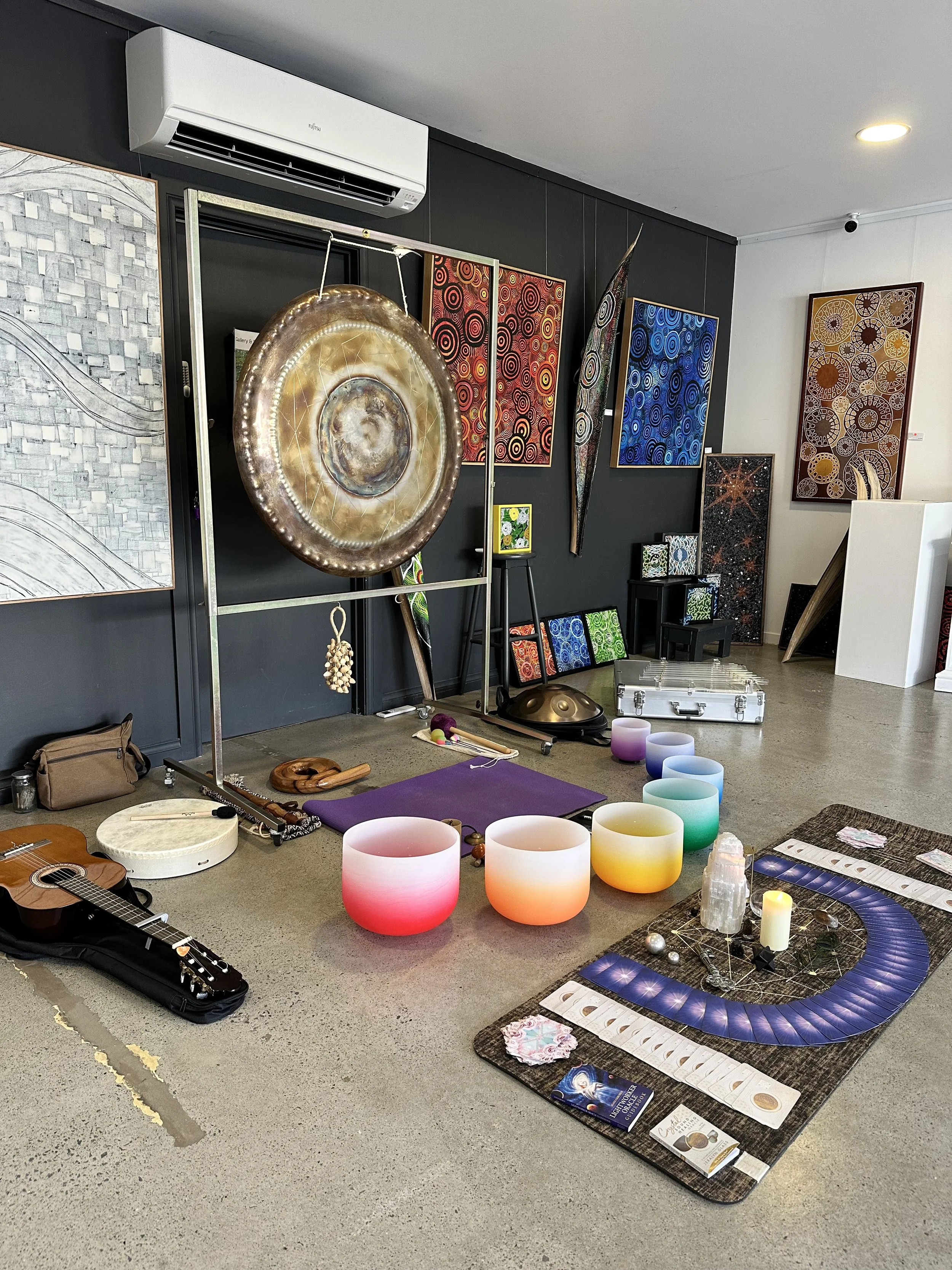 An art gallery display of colorful items including a gong, musical instruments, rainsticks, and various decorated paintings on the wall. There is also a setup of singing bowls, candles, and crystals on a mat on the floor.