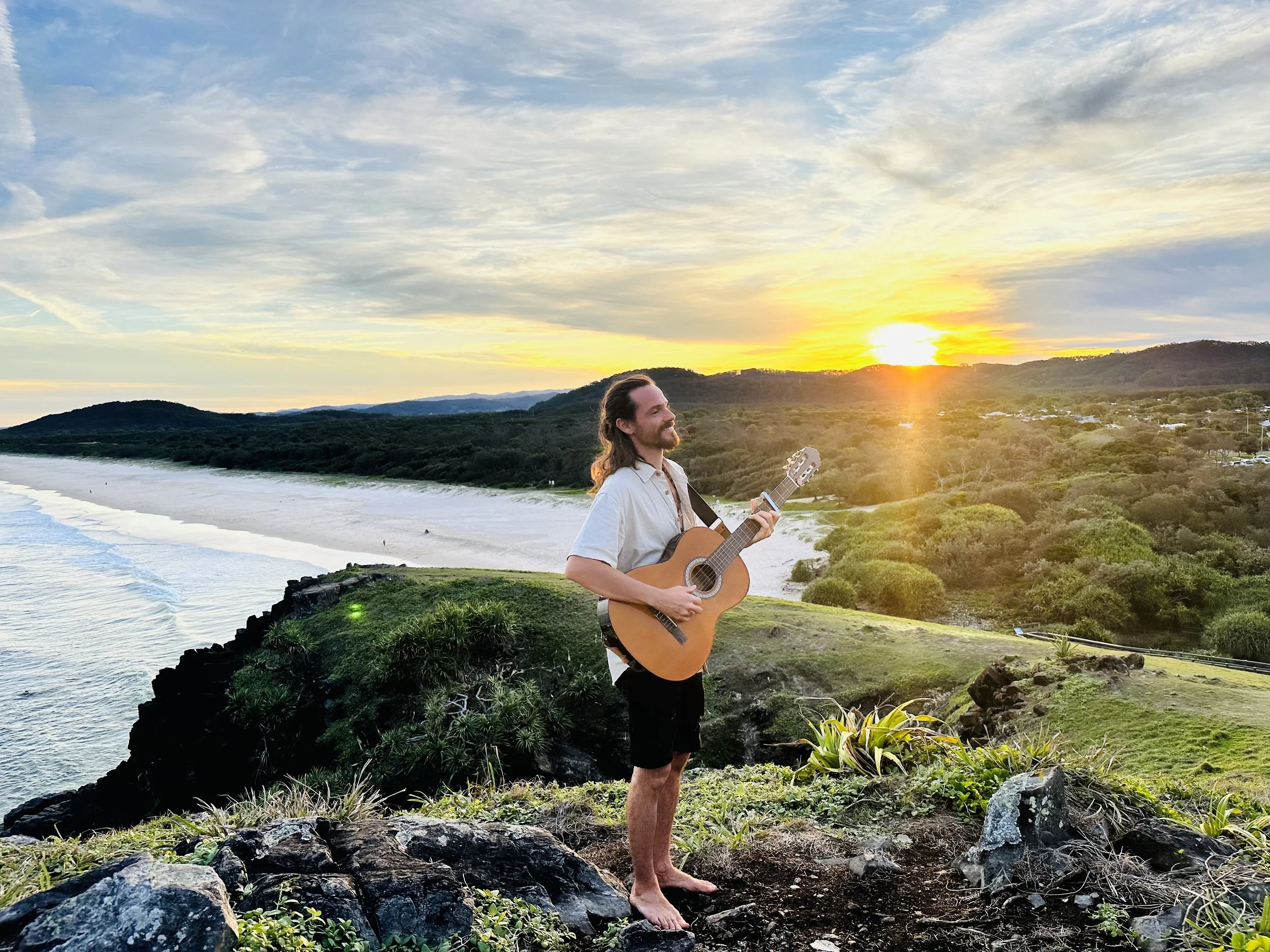 A man playing guitar on a hilltop overlooking a beach at sunset, with green vegetation and distant hills.