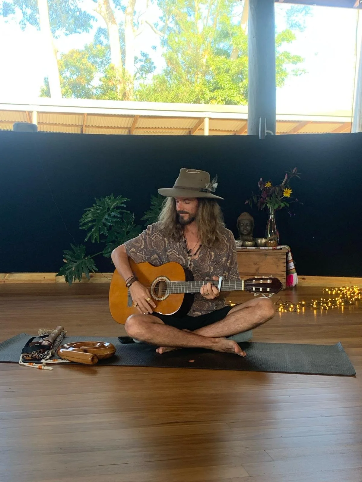 A man with long hair and a beard wearing a wide-brimmed hat, sitting cross-legged on a yoga mat, playing an acoustic guitar inside a room with wooden flooring. Behind him, there's a black wall with plants and decor, and a window showing trees and a building exterior.