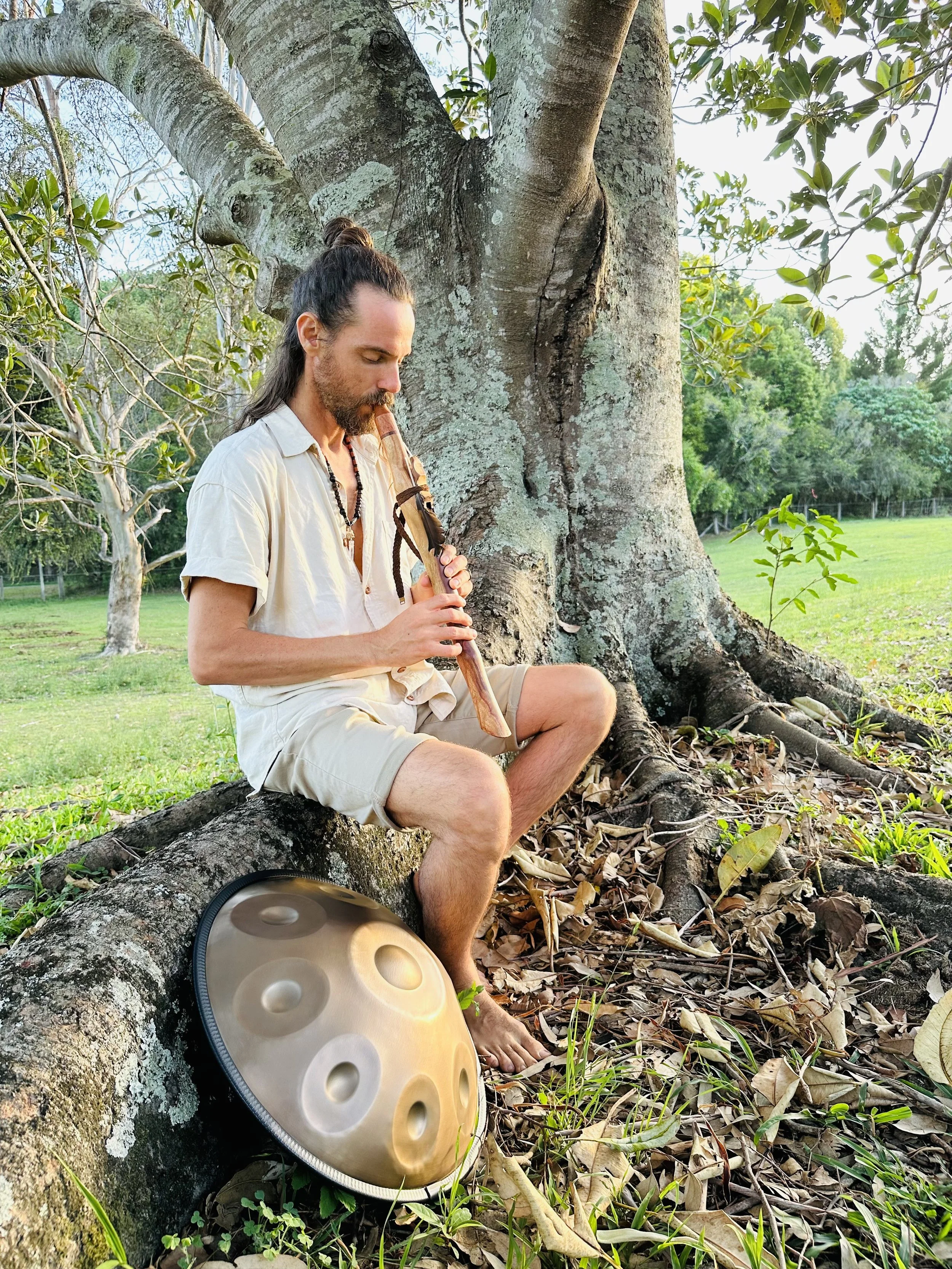 A man with long hair tied back, sitting on a fallen tree trunk in a park, playing a wooden flute. A handpan drum rests on the ground nearby. The man is dressed in light-colored casual clothing, and the background includes green trees and grass.