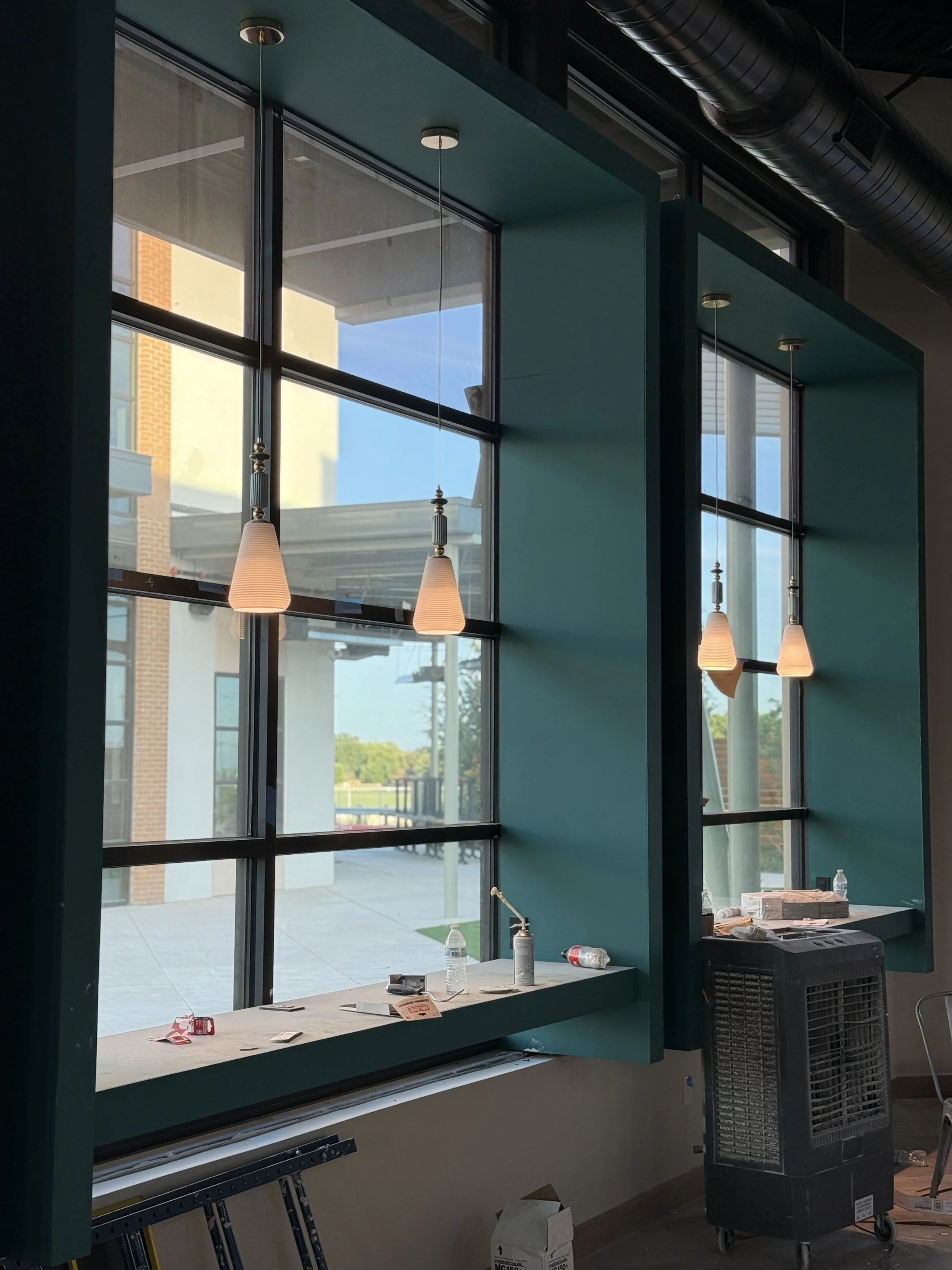 Interior of a building under construction featuring large window with a view of a courtyard outside. Three pendant lights hang from the ceiling, and there are construction materials and bottles on a windowsill.