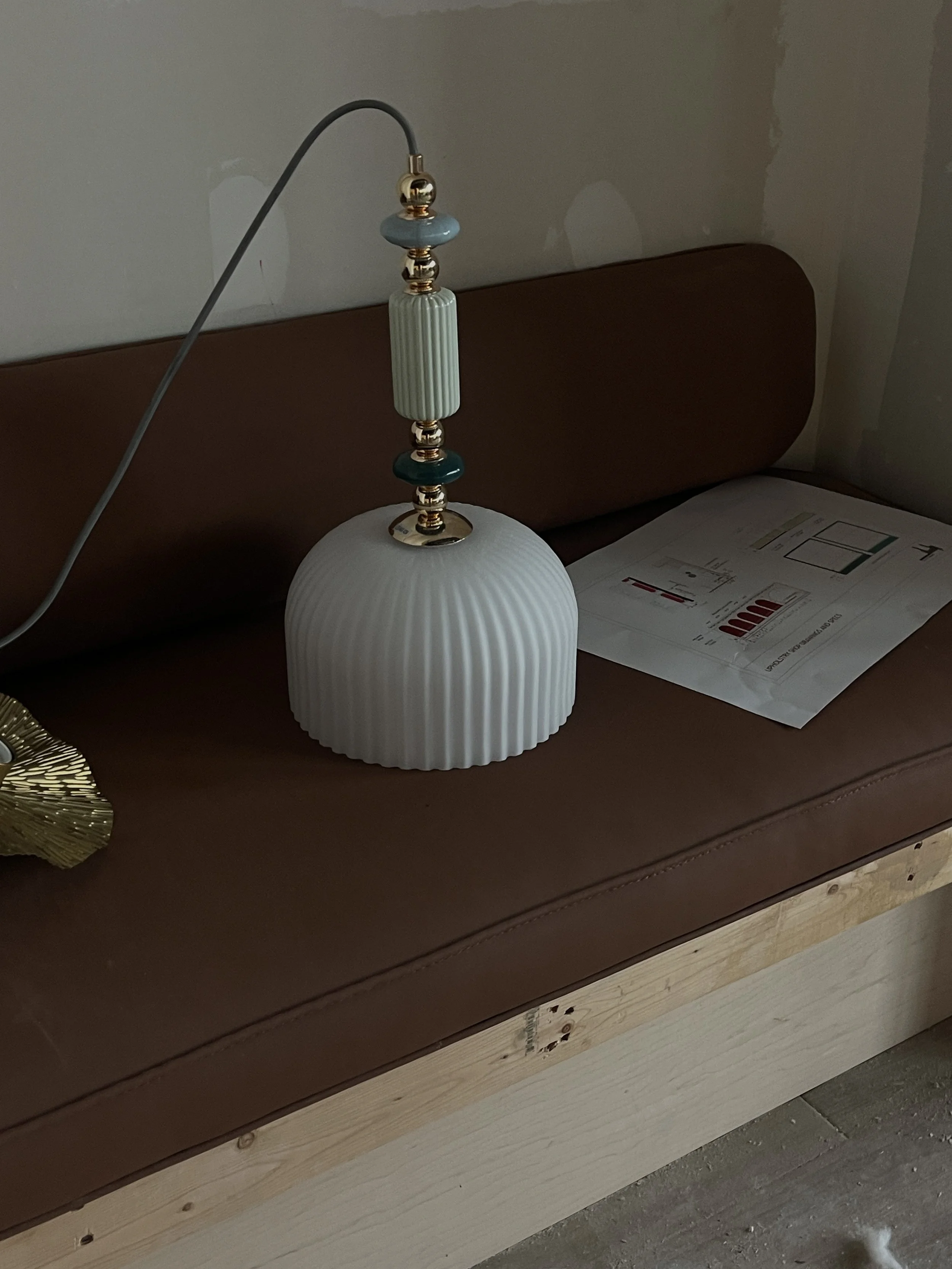 A white ceramic table lamp with a ribbed base and decorative beads on its neck. The lamp is placed on a brown leather upholstered bench with a wooden base, next to some papers or magazines.