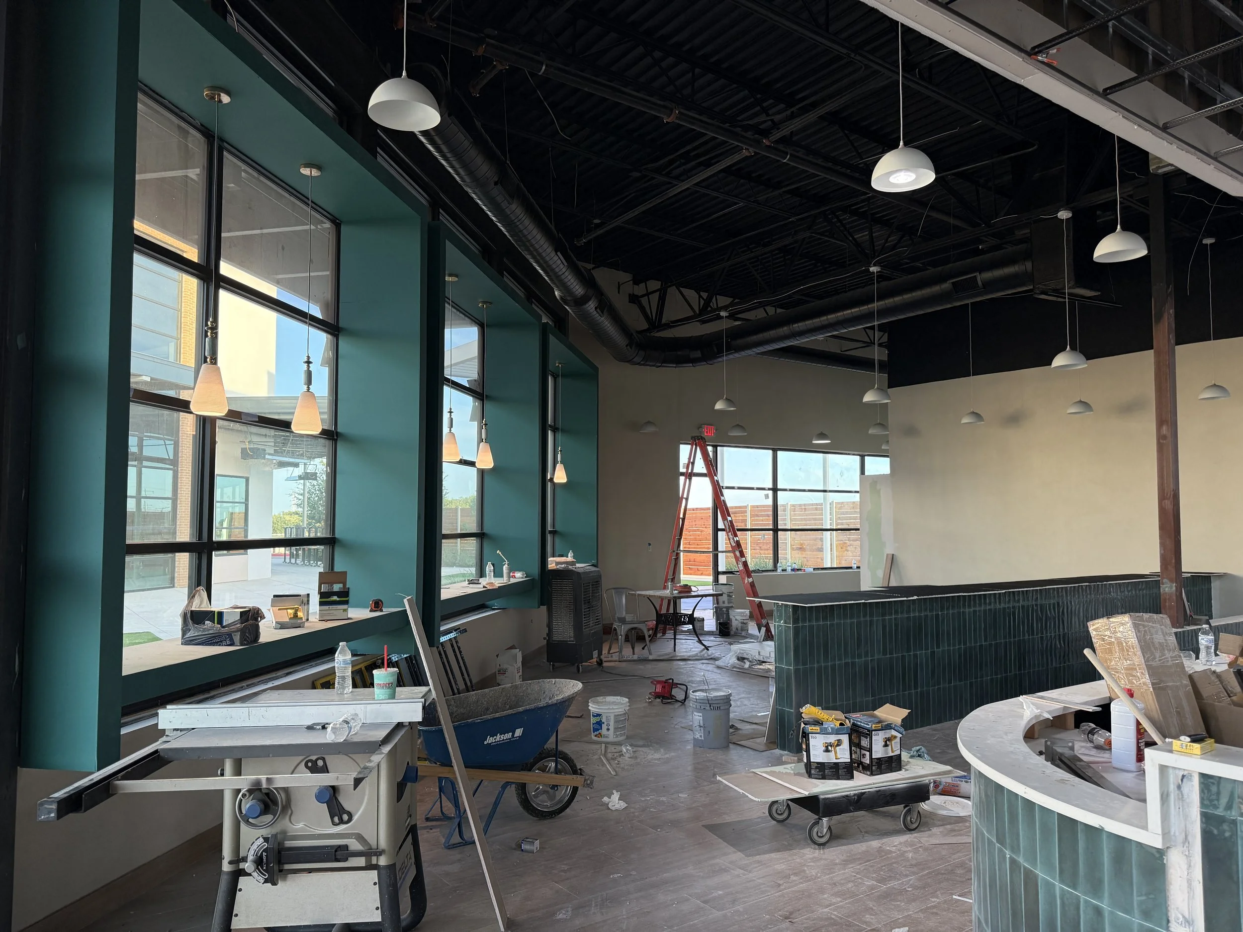 Interior of a building under construction with large windows, hanging lights, construction tools, and materials scattered around.