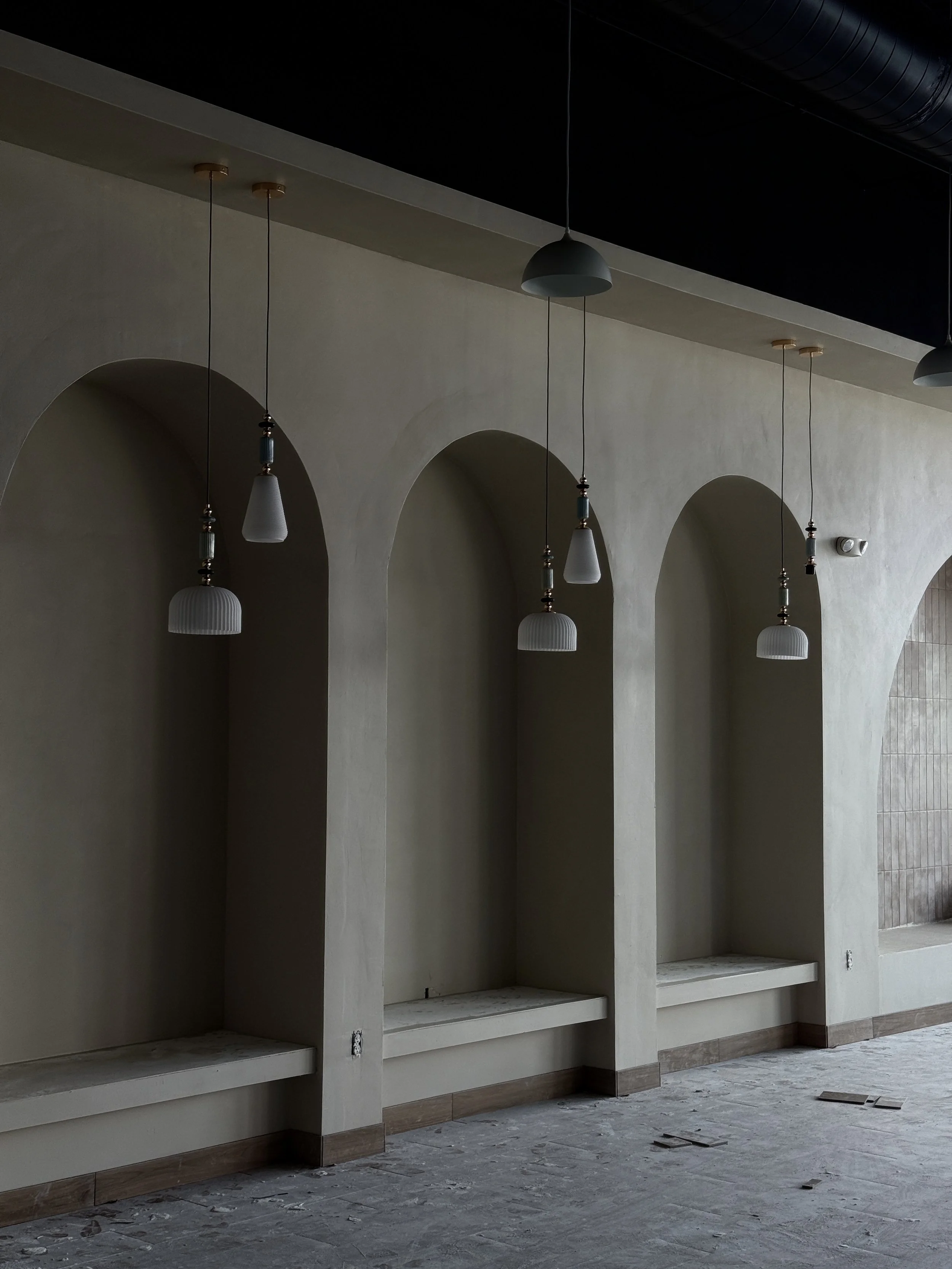 Interior of a building under construction or renovation, featuring three arch-shaped alcoves with built-in benches, hanging pendant lights, and a textured wall to the right.