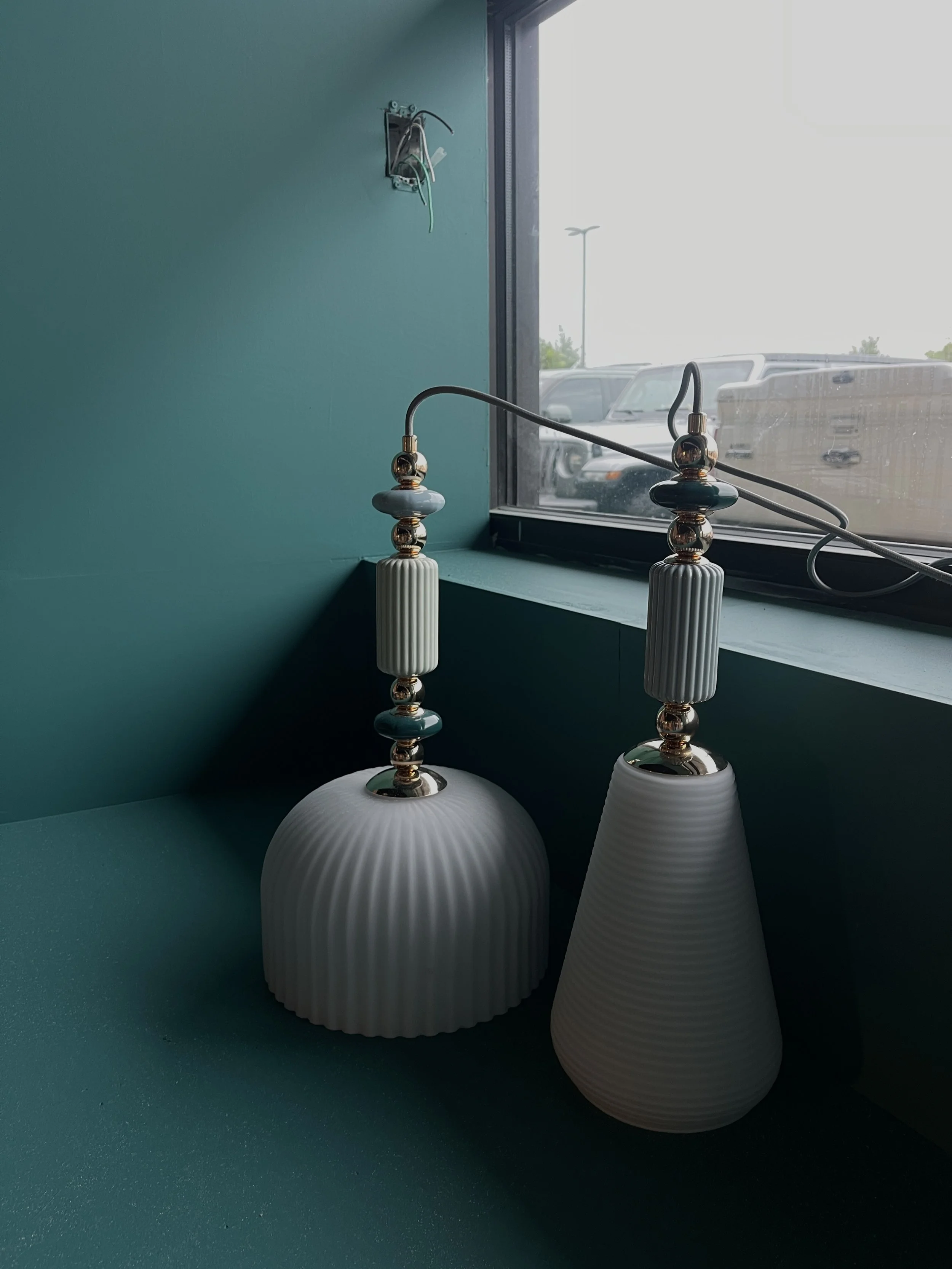 Decorative table lamps with ribbed ceramic bases and ornate metal accents, placed on a teal windowsill near an outdoor parking lot.