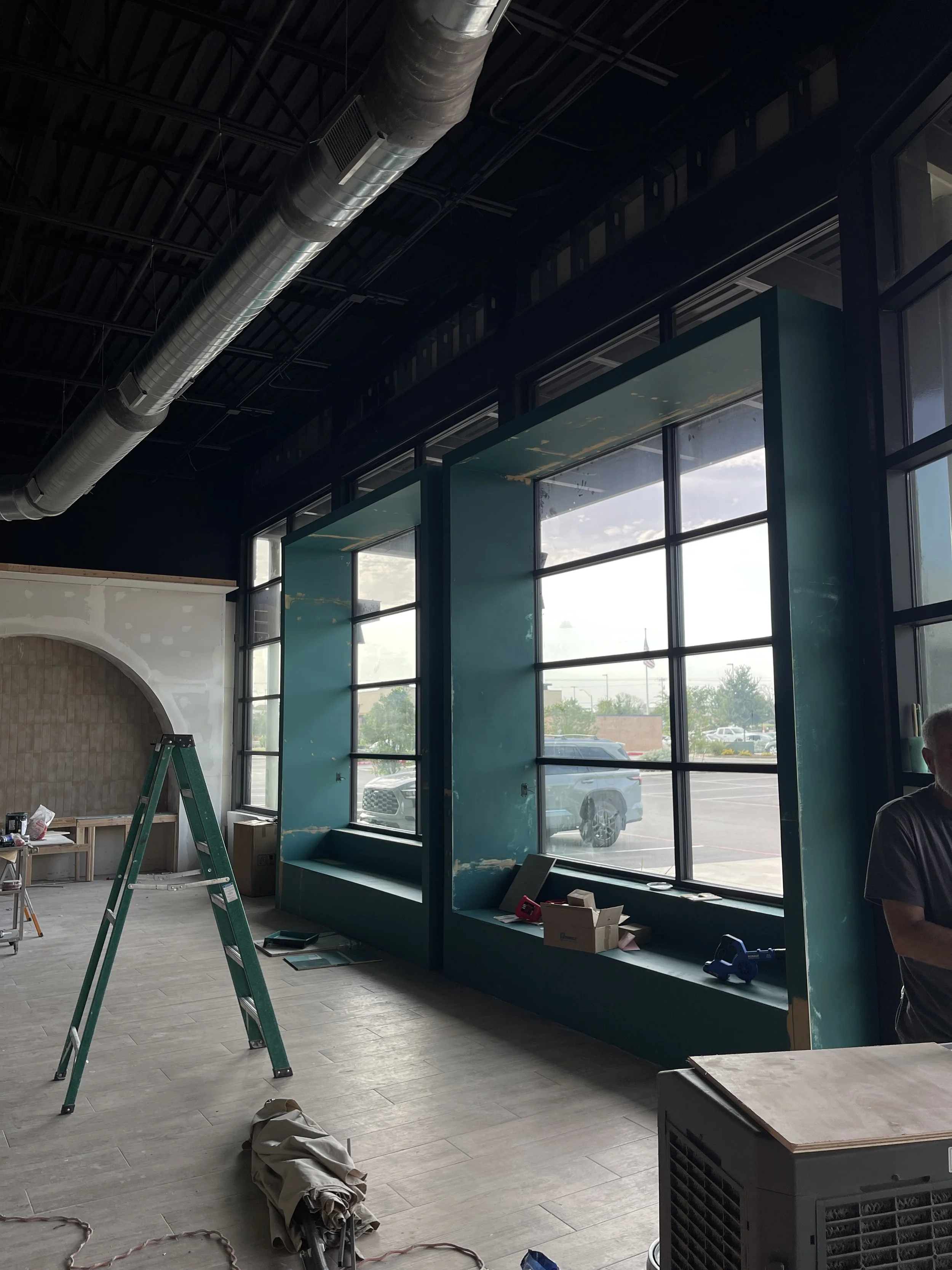 Interior of a store or cafe under construction with large windows, a ladder, and construction tools and materials.