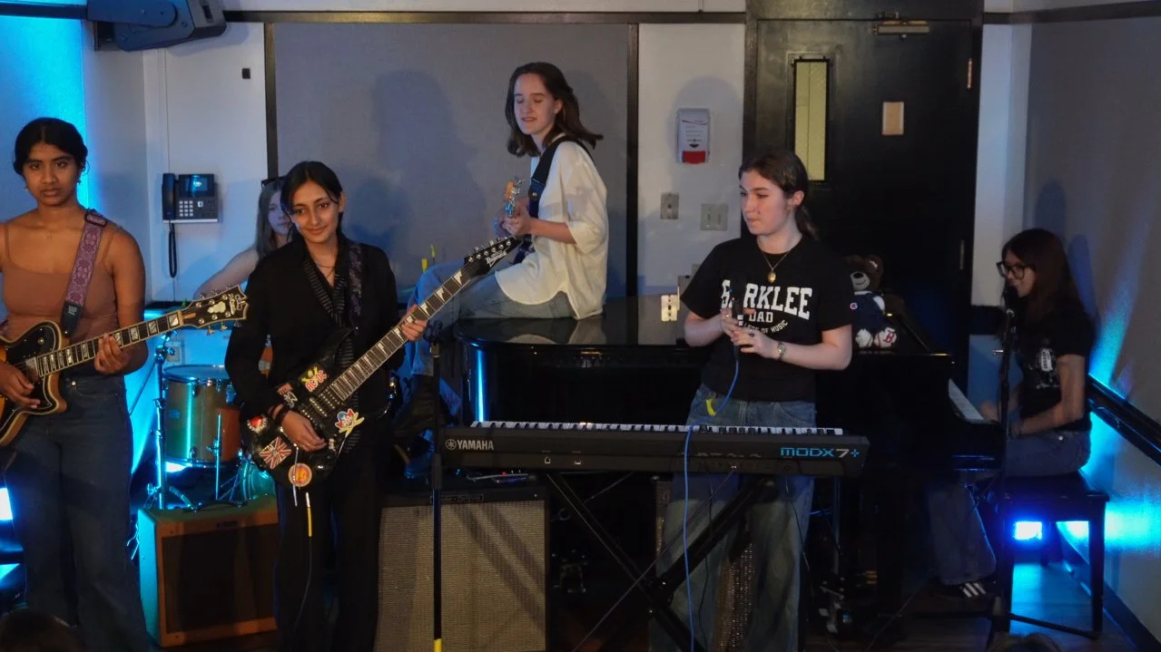 Group of young women performing music on stage with guitars, keyboard, and drums, in a room with blue and white lighting.