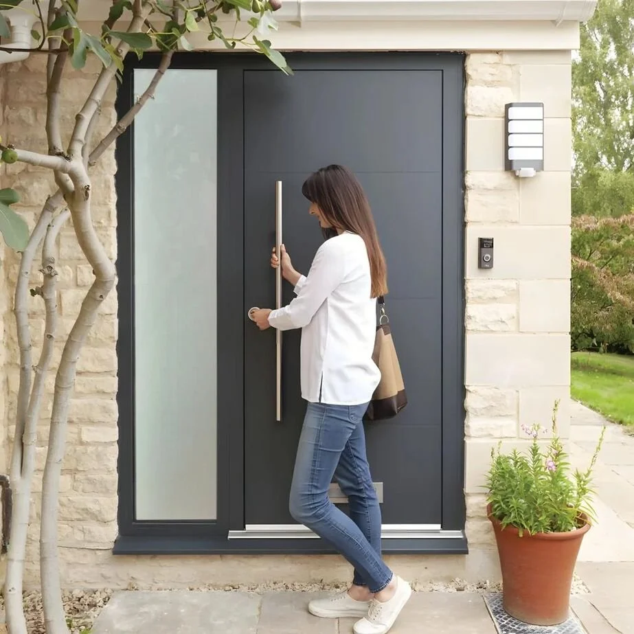 Woman wearing a white jacket and jeans opening a dark front door with a long handle, next to a potted plant and a tree