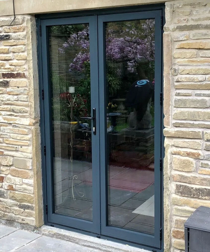 Glass double doors with black frames, similar to aluminium doors, set in a stone wall and reflecting a garden of trees and pink blossoms.