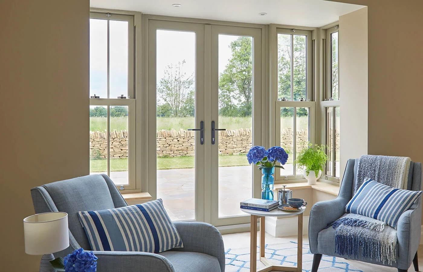 Living room with blue armchairs, blue striped pillows, side table with books and cups, vase of blue hydrangeas, two potted plants, large glass door, and windows showing outdoor patio and green yard.
