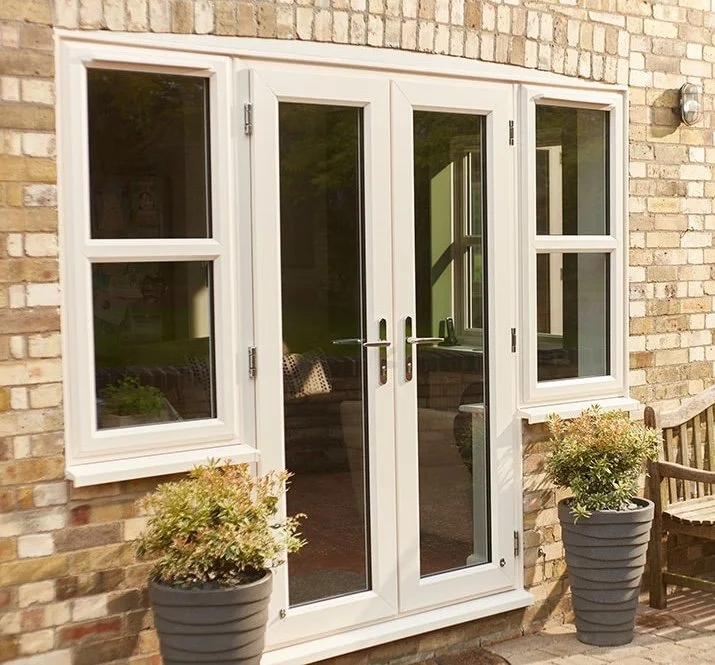 Double glass doors with white frames and black handles, surrounded by two white-framed windows on a brick wall, with potted plants on either side and a wooden bench nearby.
