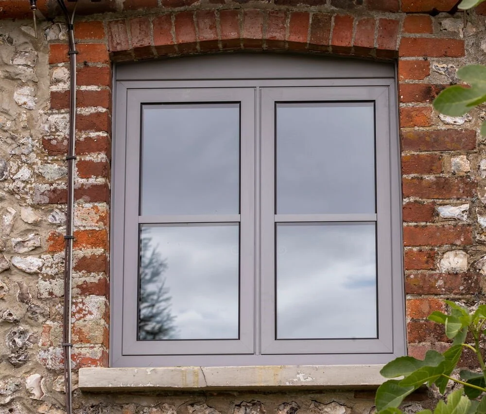 A timber bay window with grid panes reflects trees and sky, set in a brick and white wall under a tiled roof
