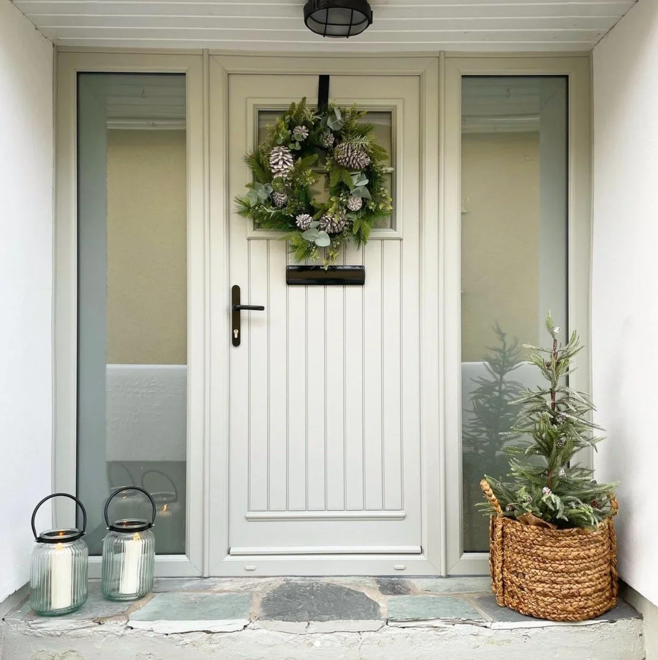 A front door decorated with a wreath, flanked by two glass side panels, with a small decorated Christmas tree in a wicker basket on the right and two lanterns with candles on the left.