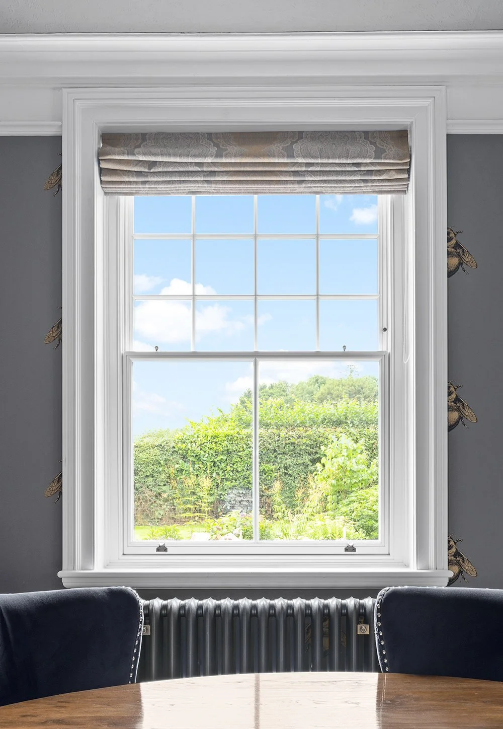 View through a large white-framed window with blinds, showing green bushes and trees outside on a sunny day.
