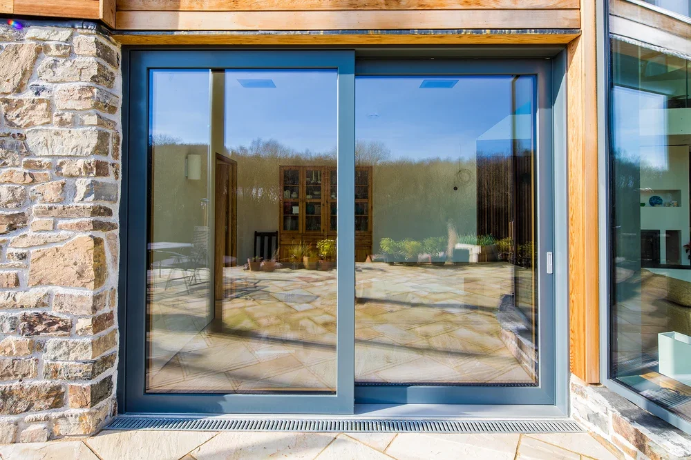 Photo of a modern house with a glass sliding door, stone and wood exterior, and a patio area visible through the door.