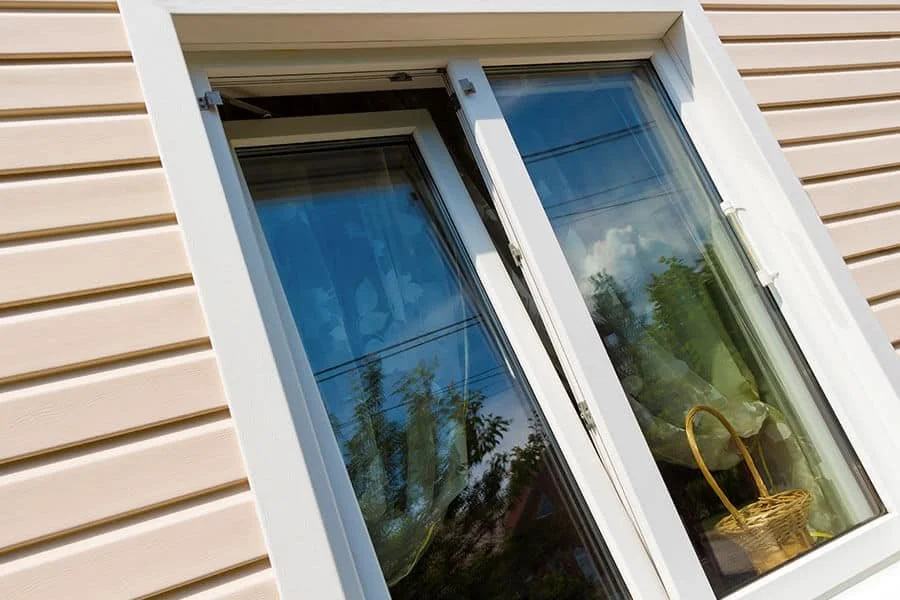 A close-up view of a window with a white frame on a house with beige horizontal siding. The window has a reflection of trees and the sky with clouds. A yellow basket is visible through the window on a windowsill inside the house.