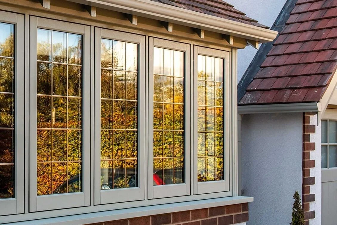 A timber bay window with grid panes reflects trees and sky, set in a brick and white wall under a tiled roof
