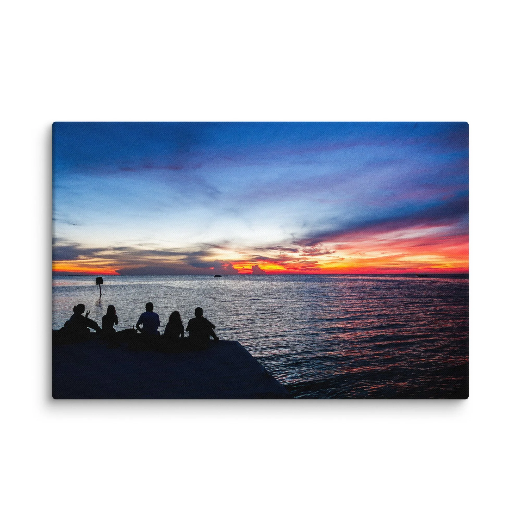 Silhouetted friends sitting on a wooden dock at sunset over turquoise Caribbean waters in Belize, with dramatic orange-pink sky and distant boat – tropical island travel photography canvas