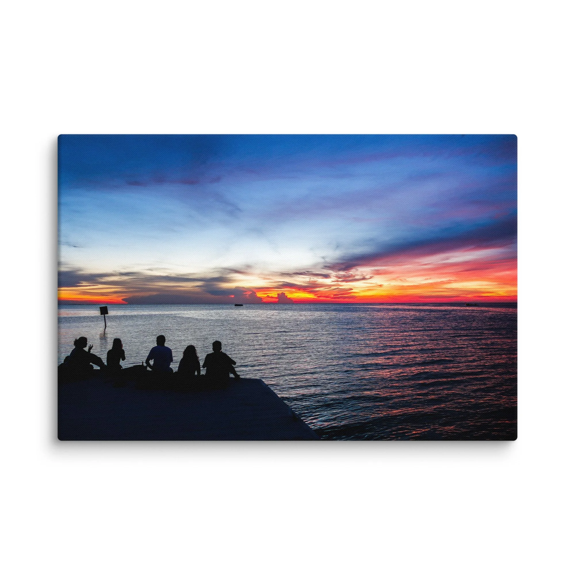 Silhouetted friends sitting on a wooden dock at sunset over turquoise Caribbean waters in Belize, with dramatic orange-pink sky and distant boat – tropical island travel photography canvas
