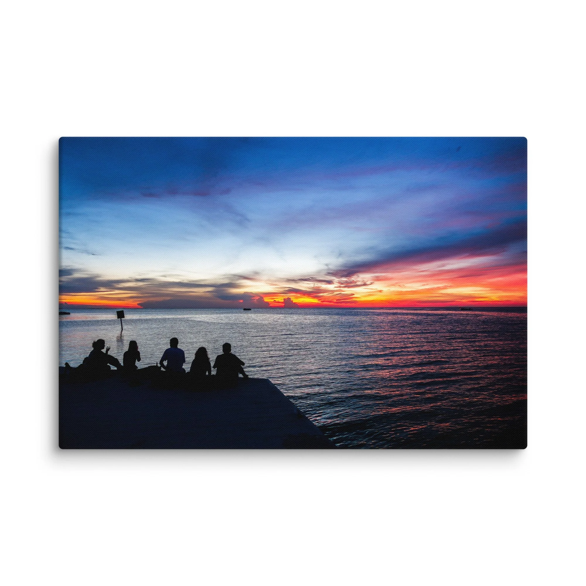 Silhouetted friends sitting on a wooden dock at sunset over turquoise Caribbean waters in Belize, with dramatic orange-pink sky and distant boat – tropical island travel photography canvas