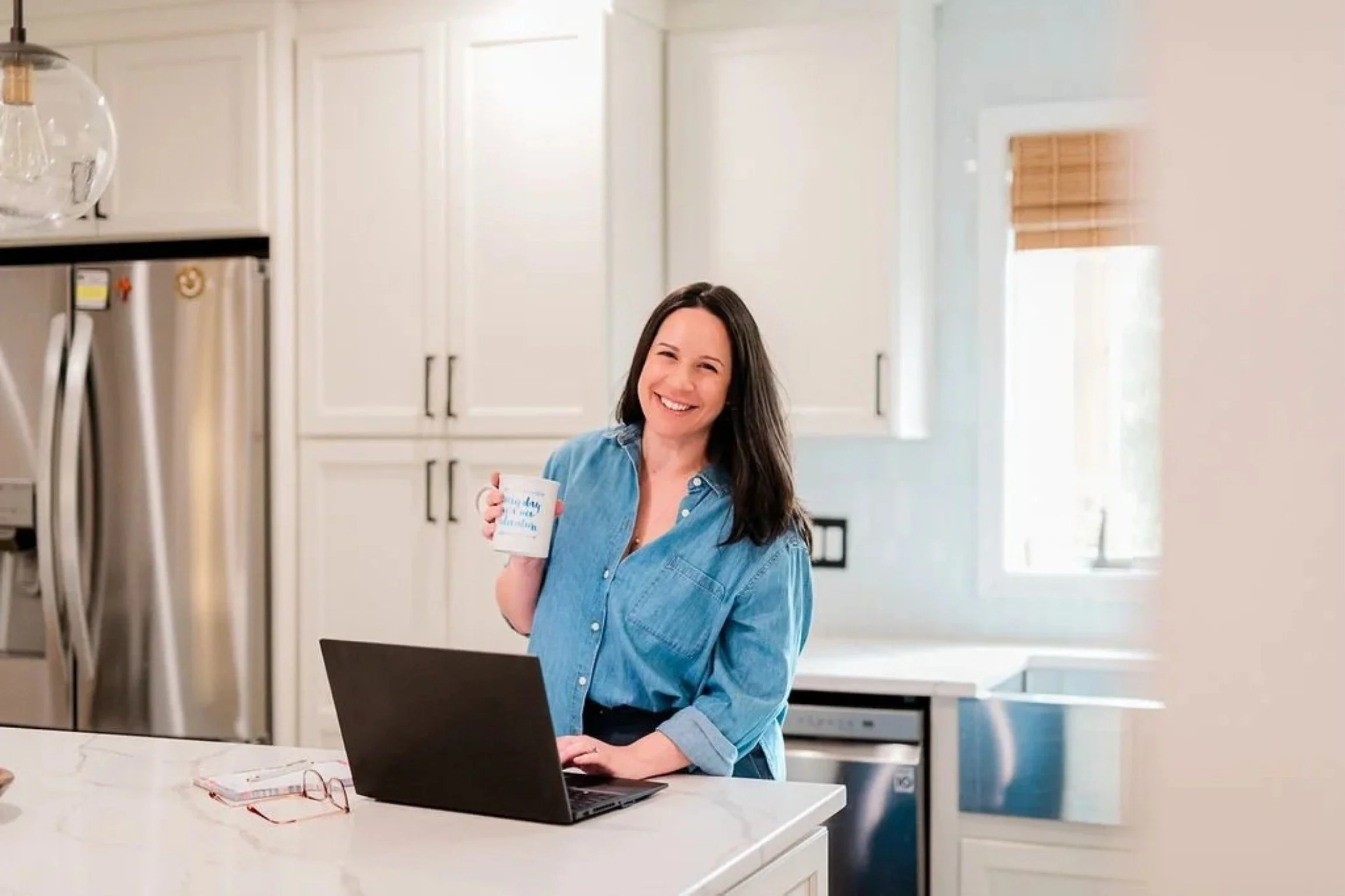 Angela, website and brand designer, stands in a white kitchen holding a coffee mug wearing a chambray shirt and working on a client's website on her kitchen island