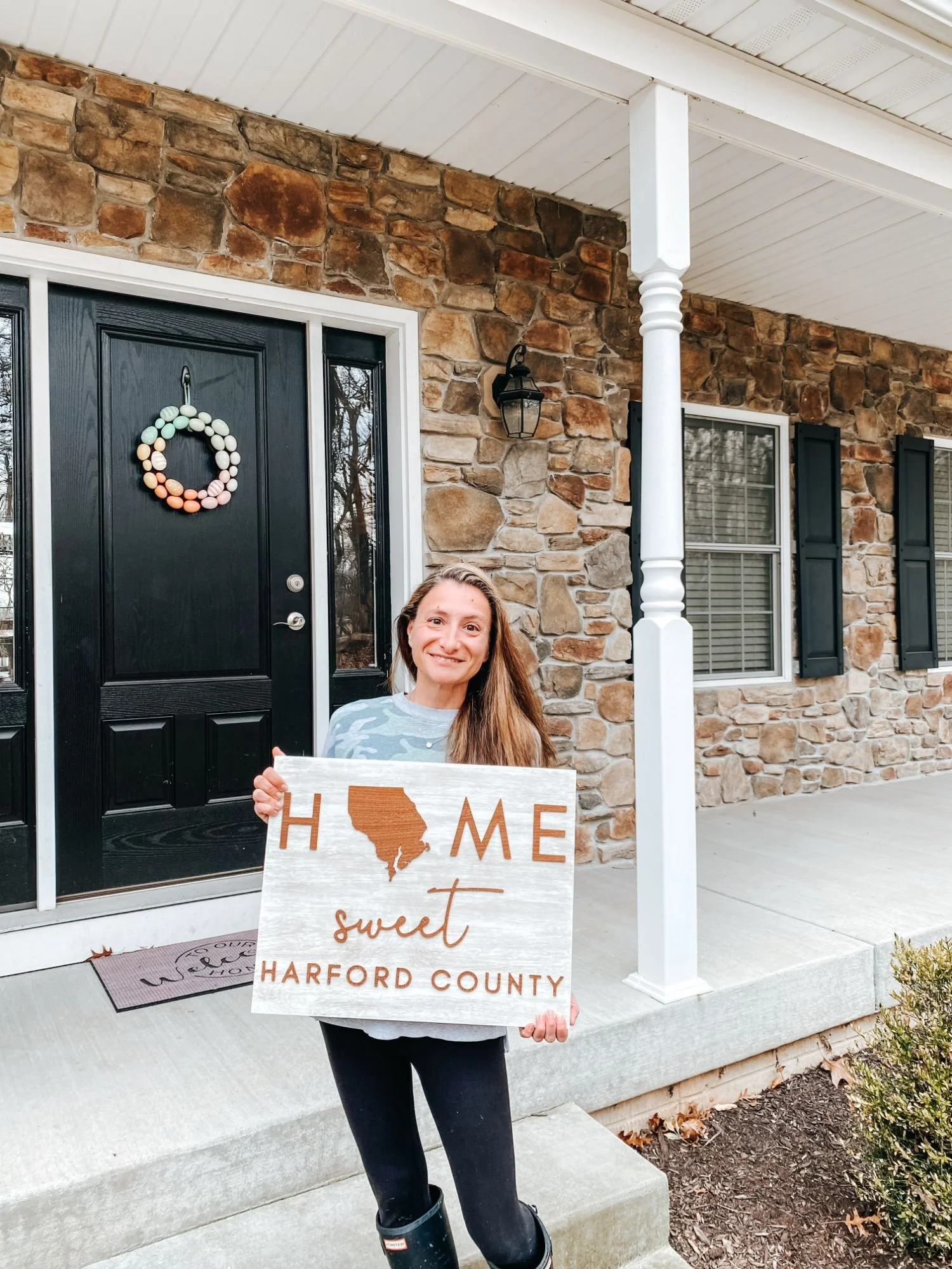 Ange, The Harford County Mom, stands in front of a house holding a "Home Sweet Harford County" sign