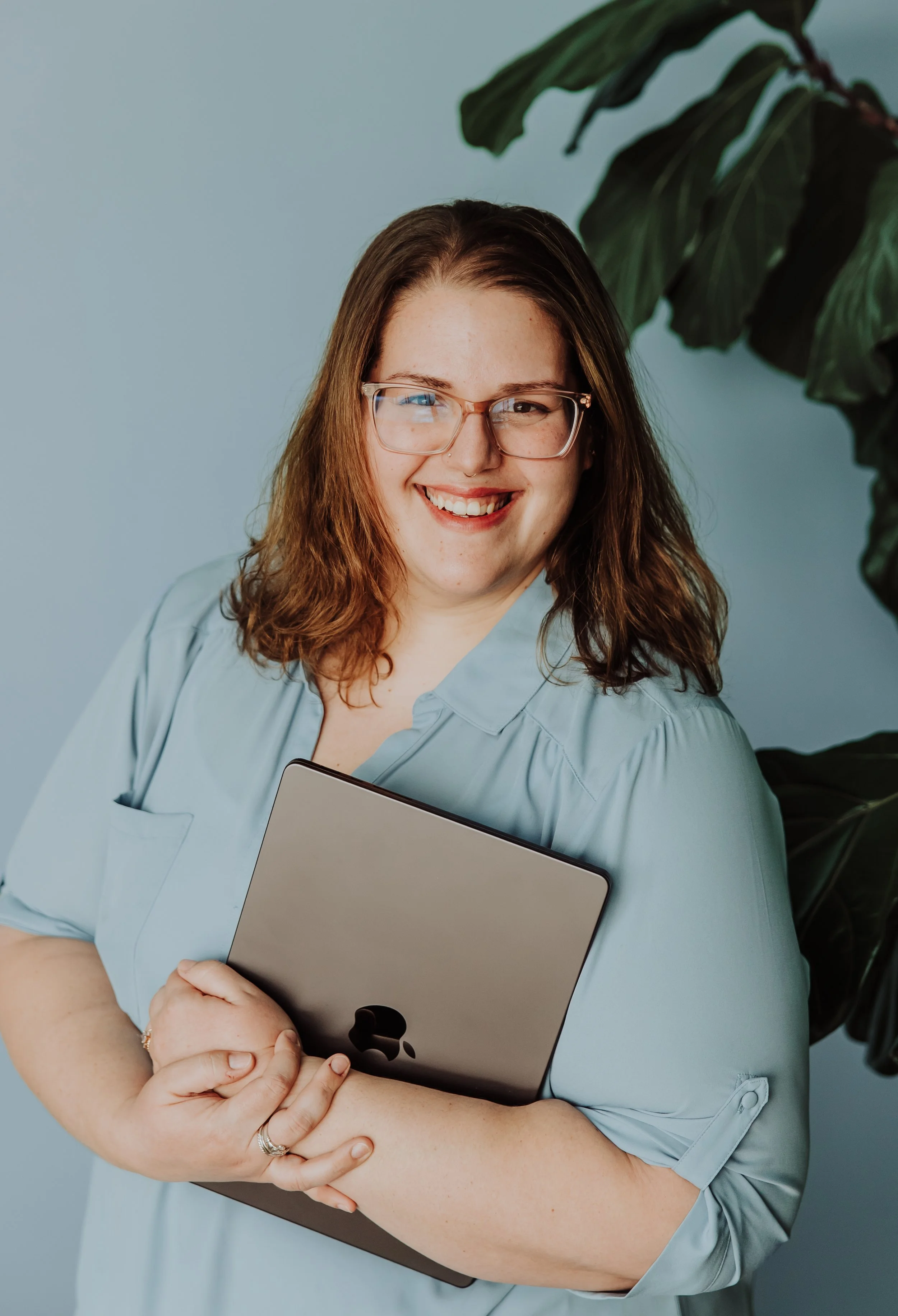 A woman with glasses holding a closed laptop and smiling at the camera, standing in front of a light blue wall with a large green plant partially visible on the right side.