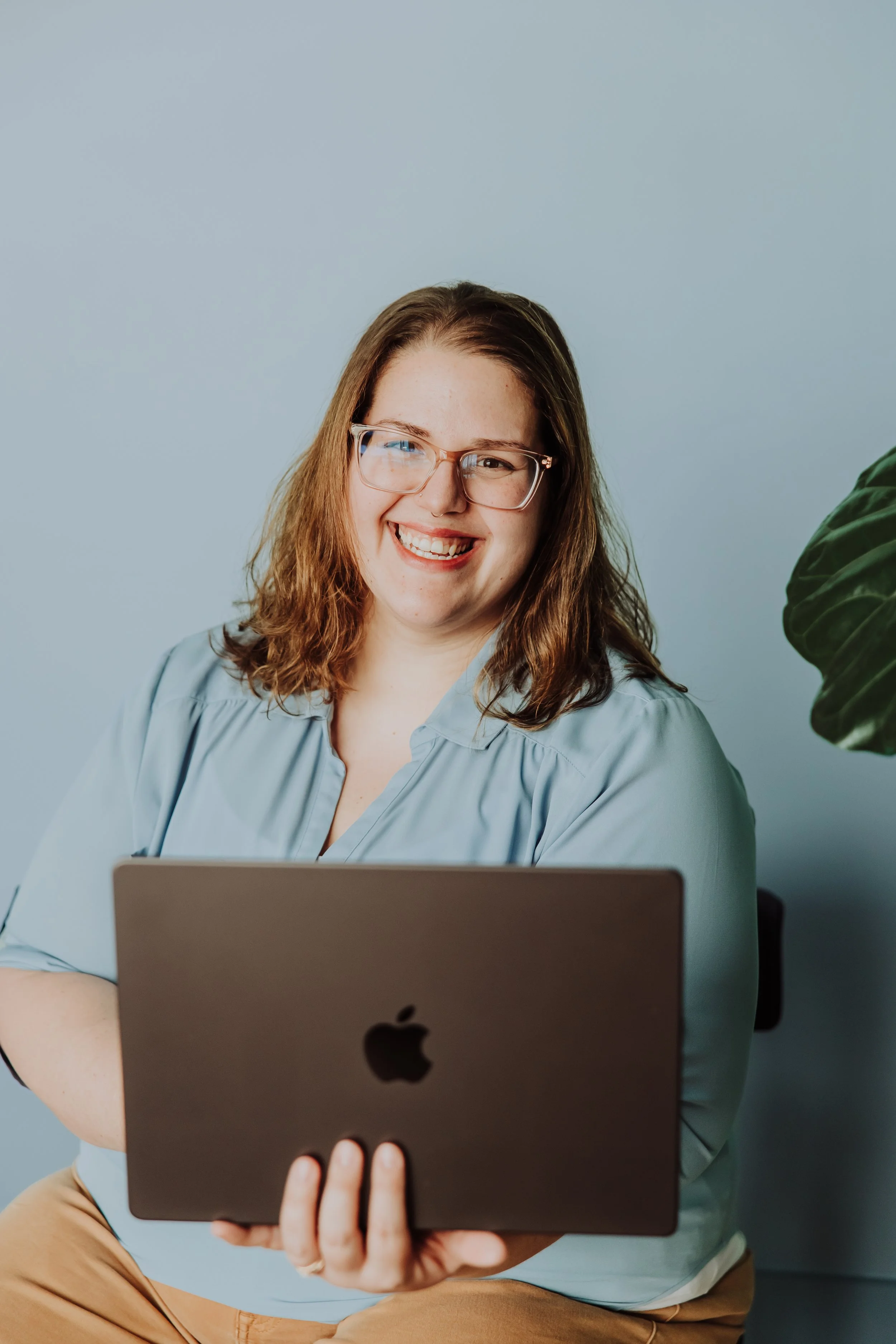 A smiling woman with glasses holding a closed MacBook in front of her, sitting by a large green plant. She has shoulder-length hair and is wearing a light blue blouse.