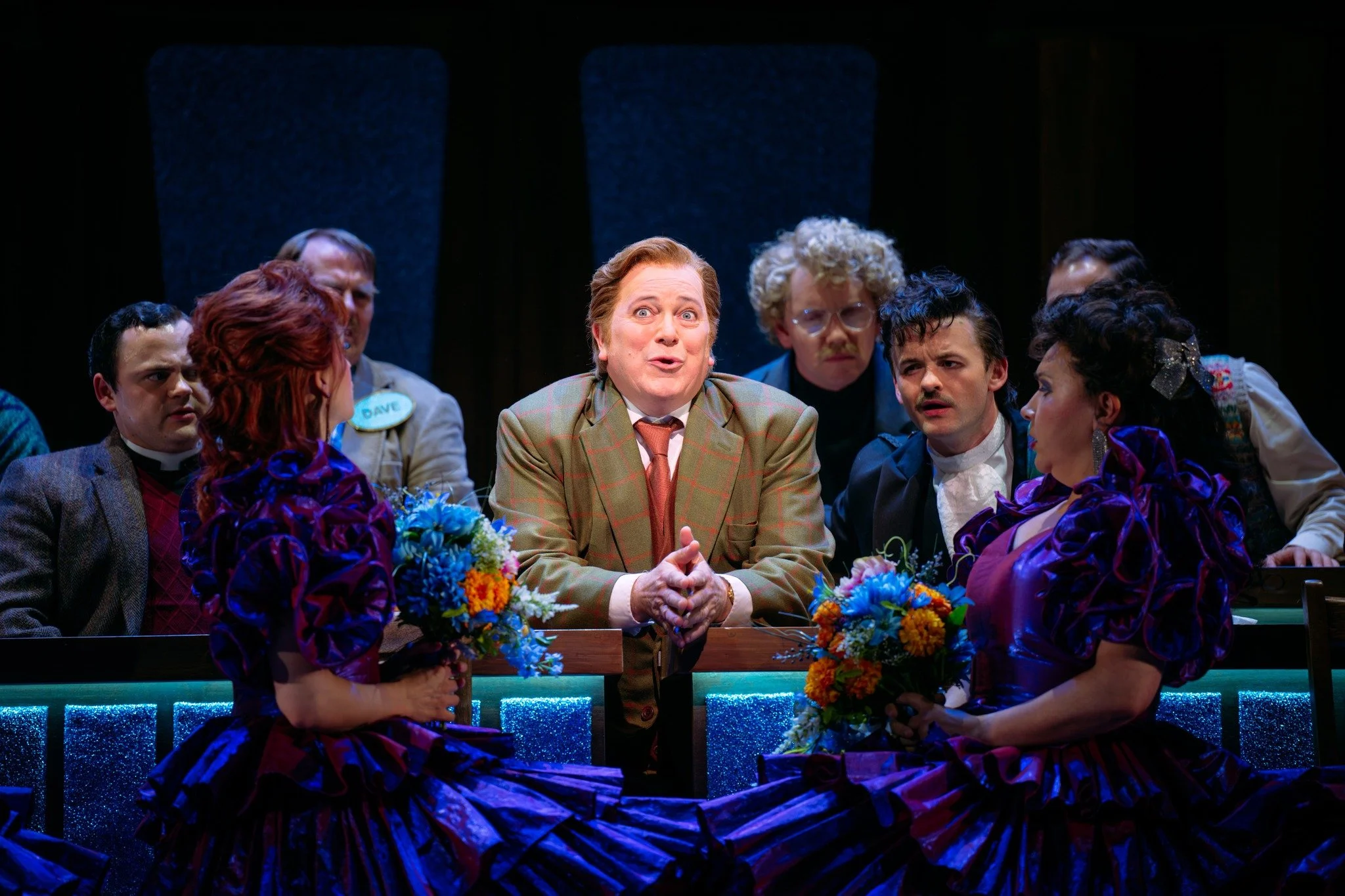 People gathered around a table on stage during a theatrical performance, with two women in purple dresses holding bouquets of flowers, and a man in front speaking or singing, with others listening attentively.