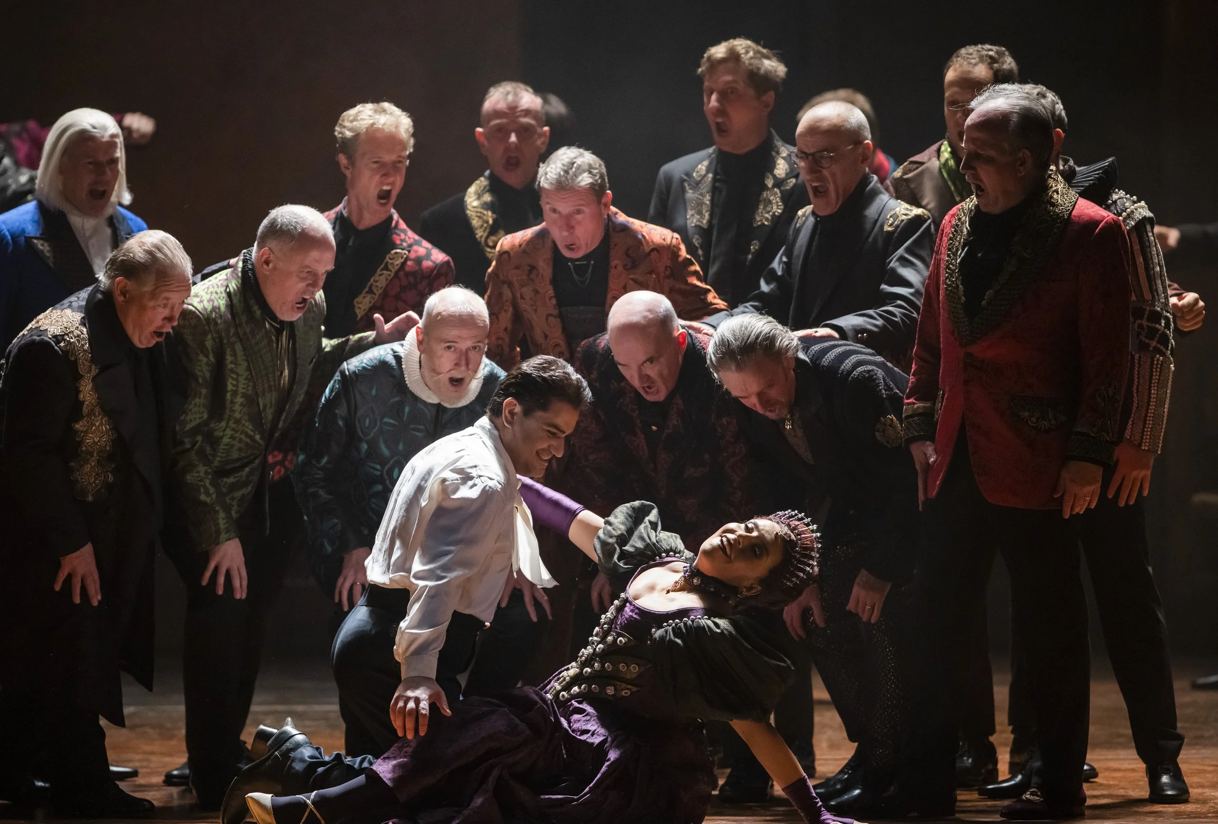 The scene shows a woman in a dark costume lying on the floor, surrounded by a group of men in colourful, period-style costumes, all appearing angry or shouting.