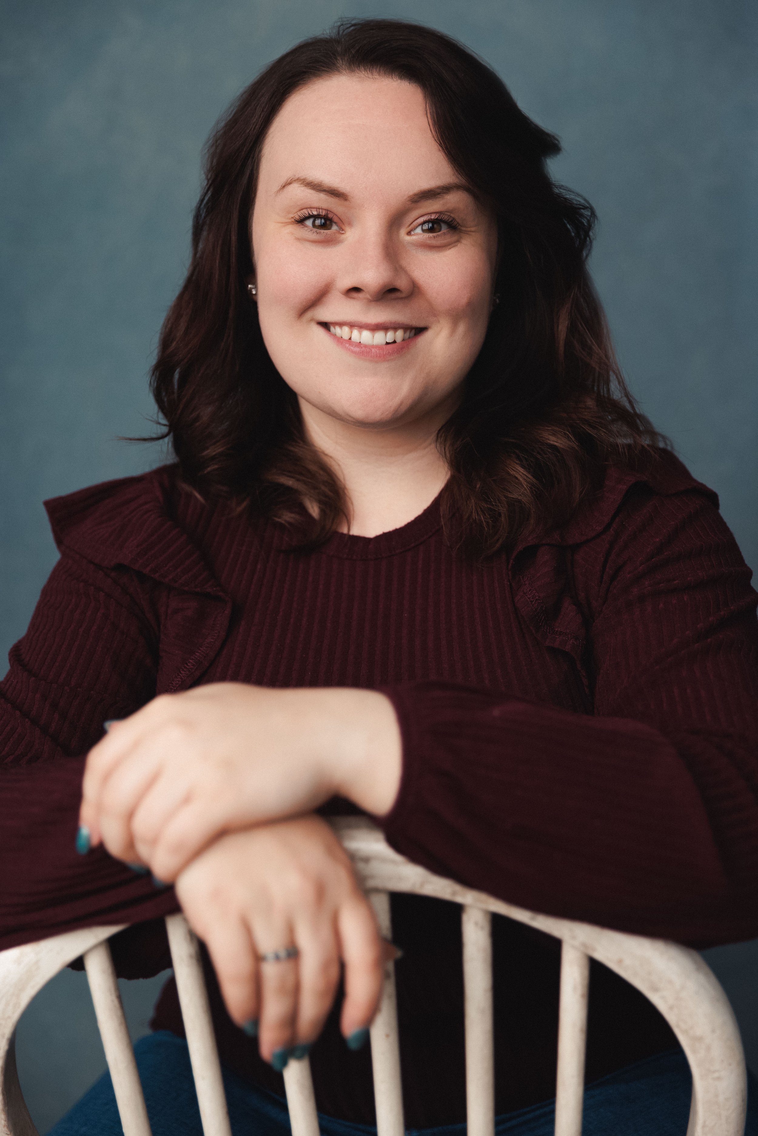 A woman with shoulder-length dark brown hair, smiling, wearing a burgundy ribbed sweater, sitting with her arms resting on a white chair against a blue background.