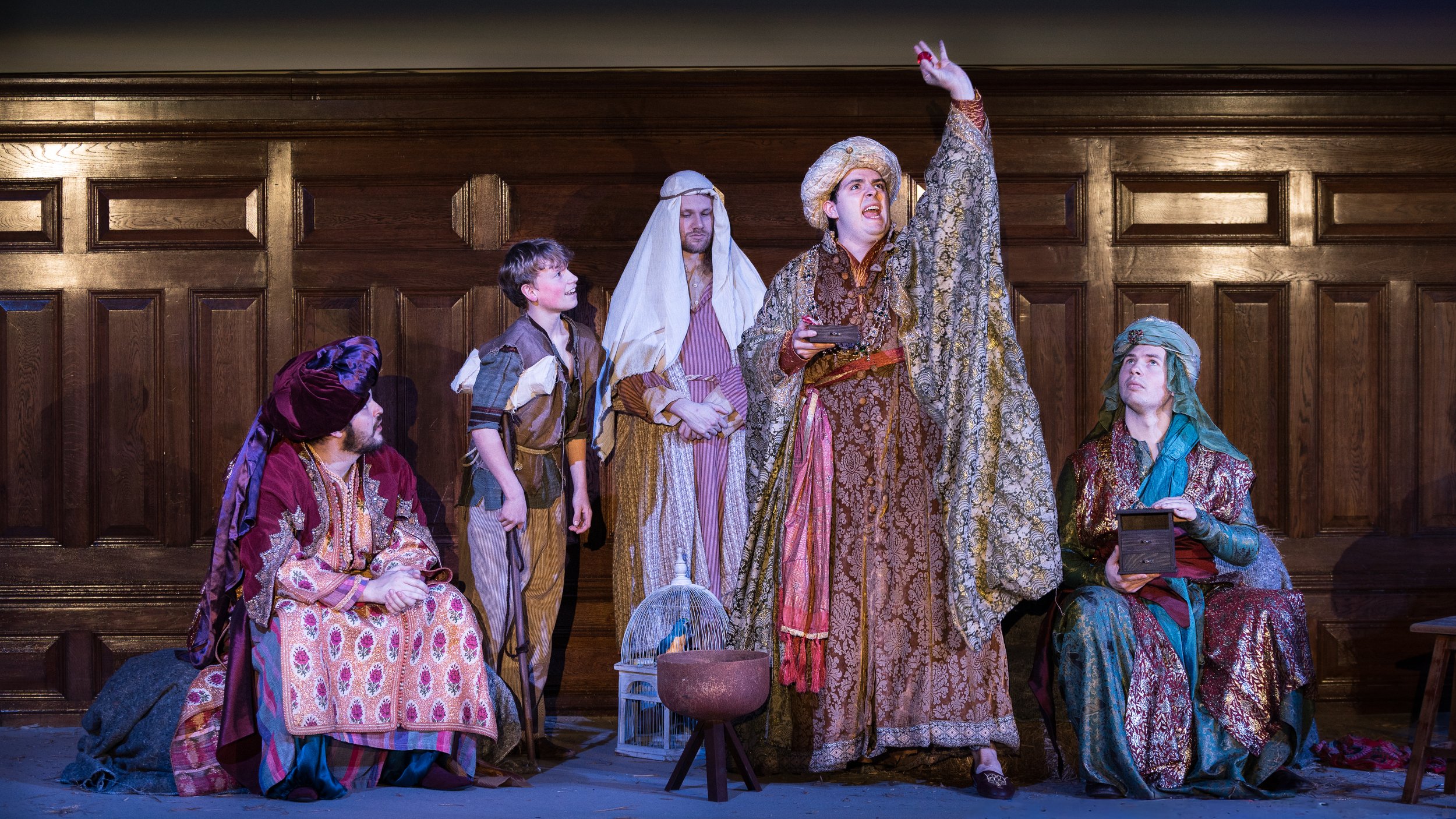 Three men in colourful, elaborate patterned costumes, a servant in plain clothes, and a young boy dressed in rags, pose on a stage in a theatrical scene, with one standing and raising a red ruby upwards in his left hand while the others sit or stand 