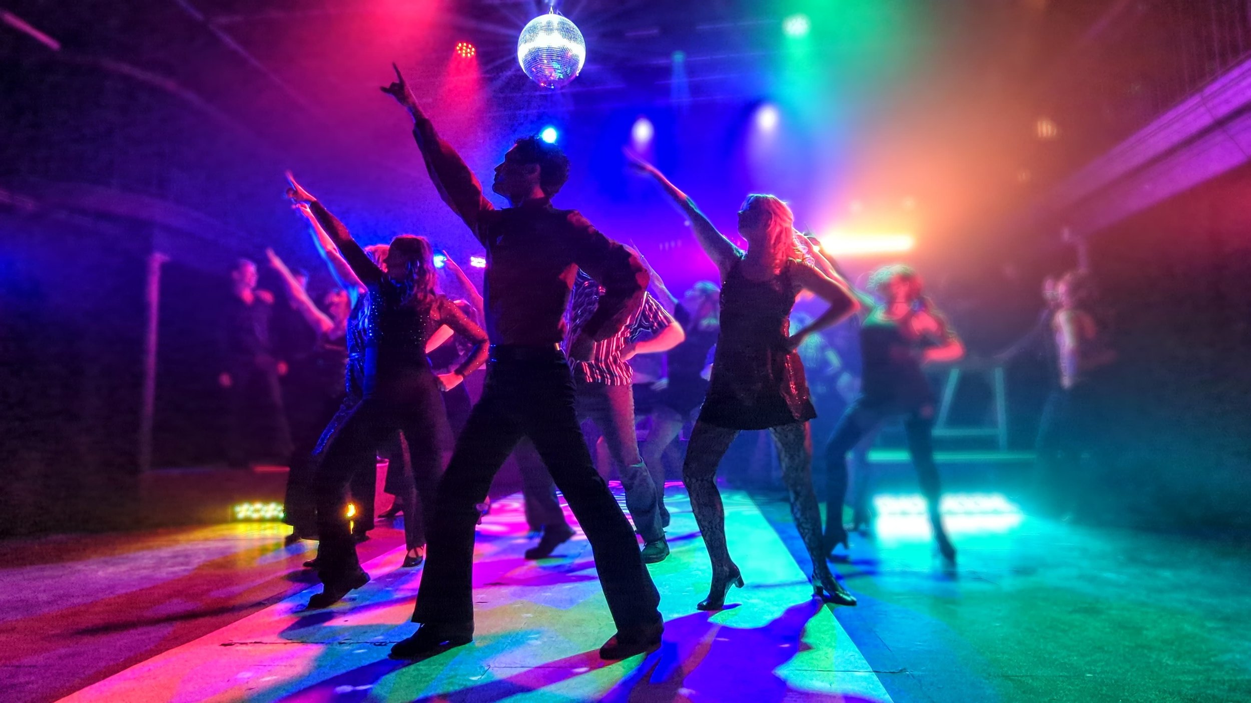 People dancing energetically under a disco ball with colourful lights on a vibrant dance floor, creating a lively and joyful atmosphere.