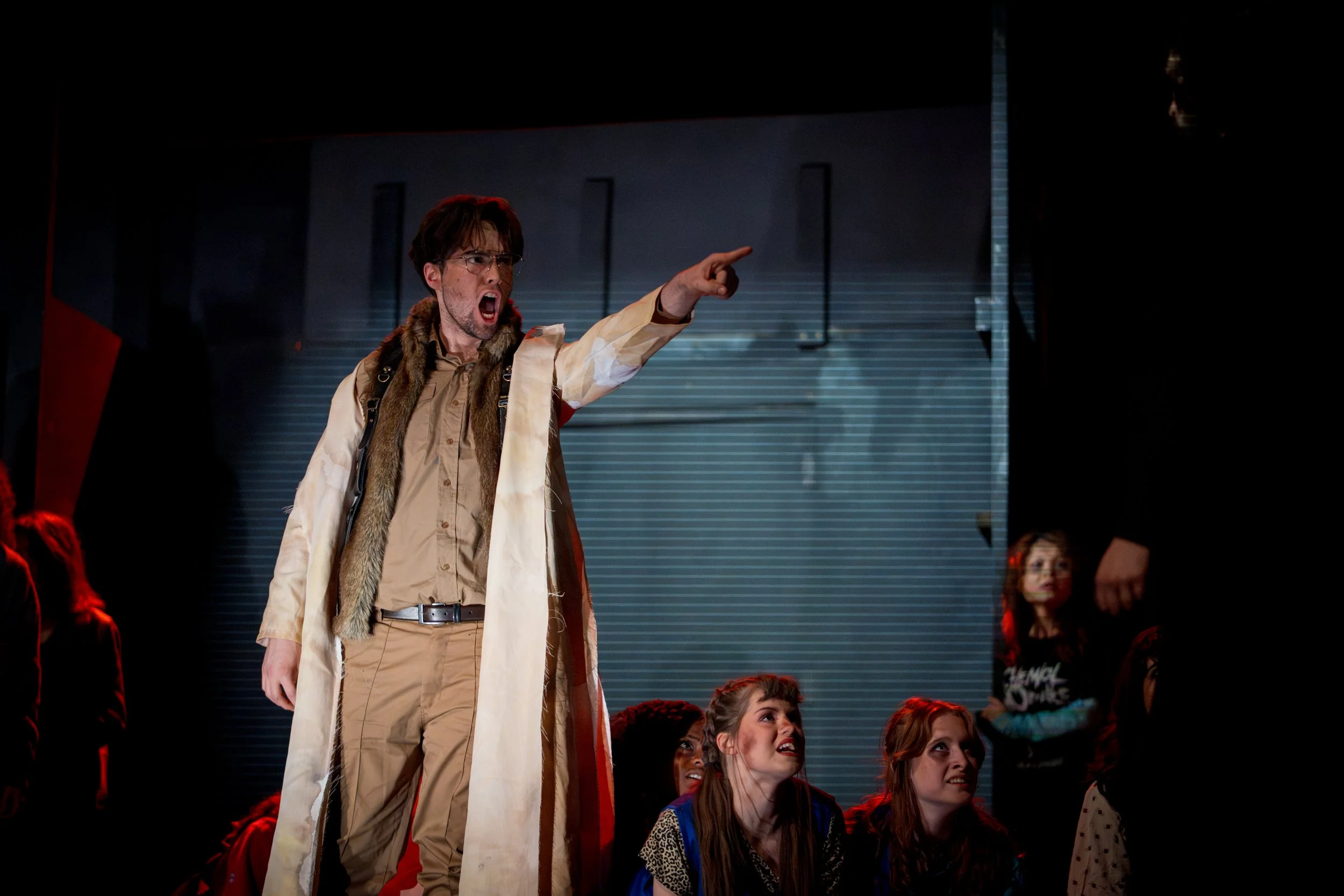 A man wearing a long coat, stands and passionately points with his left hand, while people sitting below watch in awe.