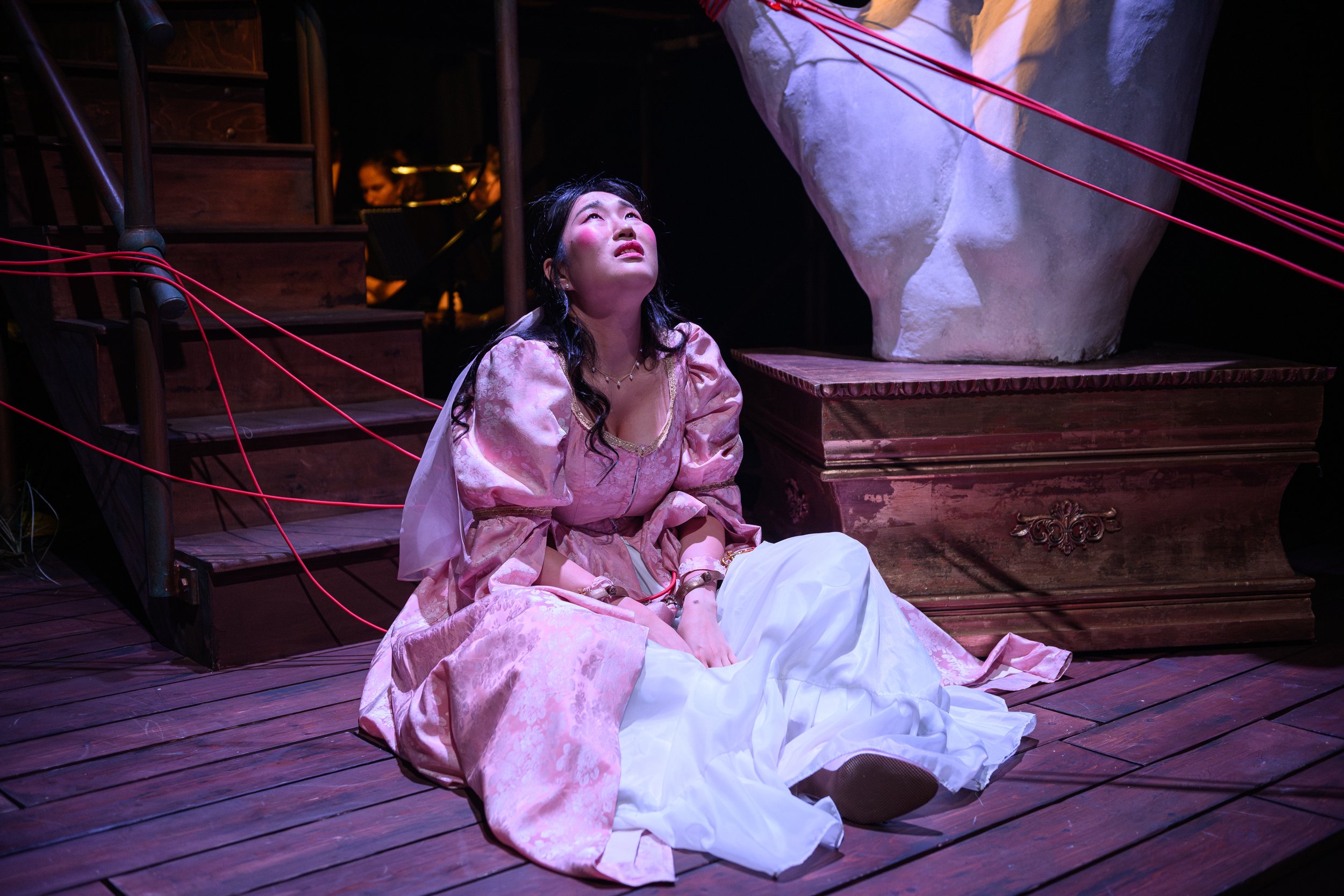 A woman dressed in a pink and white costume sits on a wooden stage floor, looking upward with a hopeful or pleading expression. She is surrounded by theatrical props, including a large white sculpture and a staircase with red ropes.
