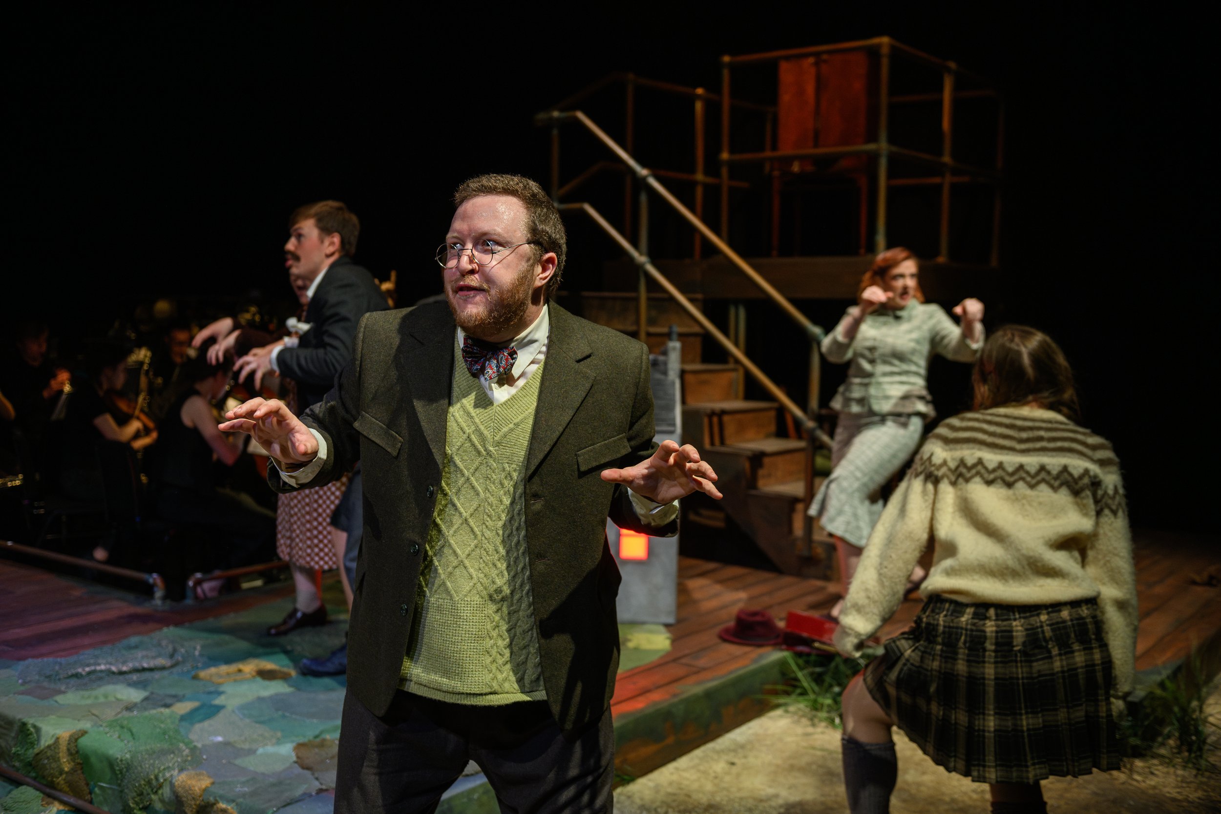 Actors in vintage attire perform energetically onstage, with hands raised and bent at the wrists, and expressions of surprise. A staircase and dim lighting set an atmospheric tone.