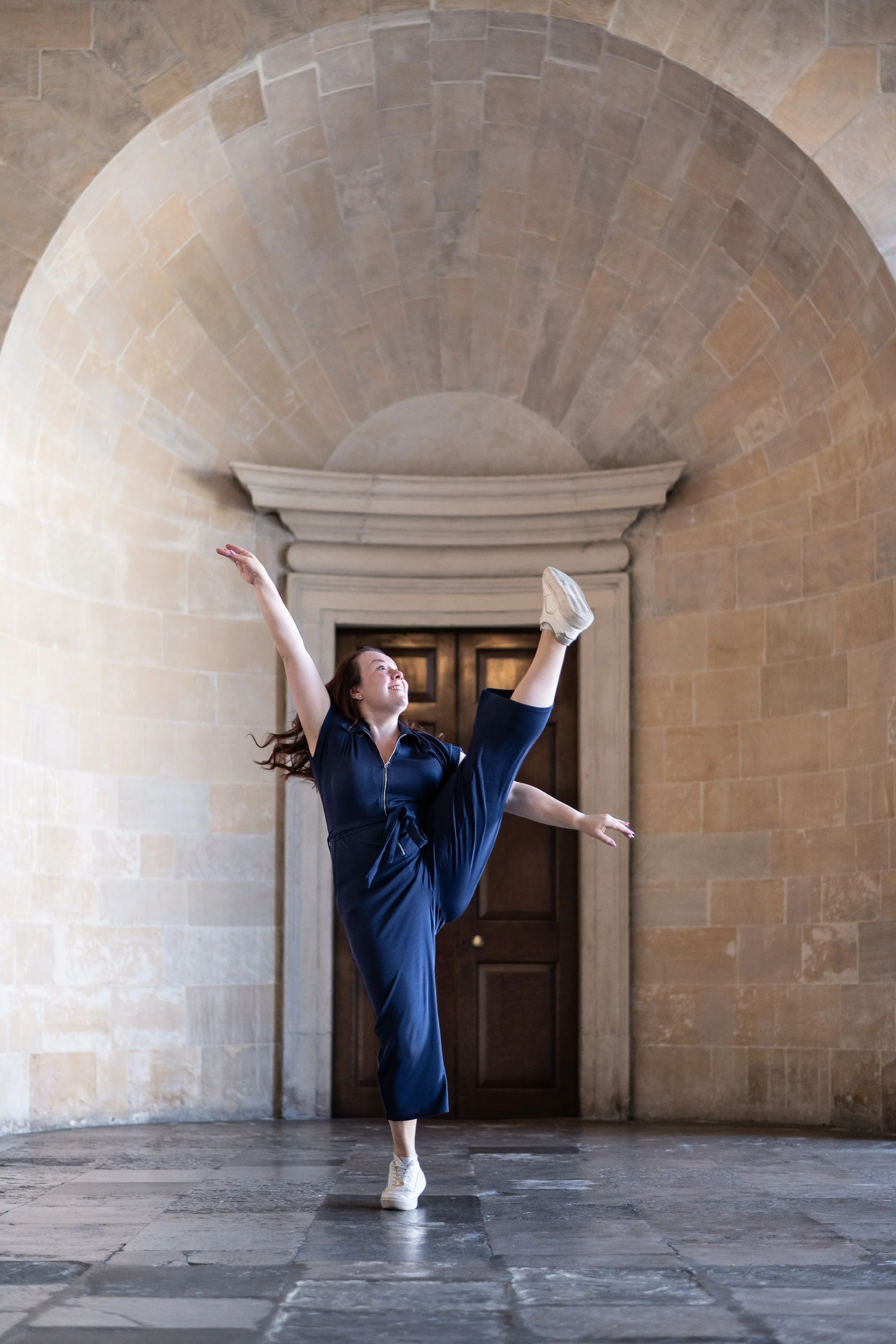 A woman in a navy jumpsuit performing a dance pose with one leg raised high and arms extended, in a stone archway corridor.