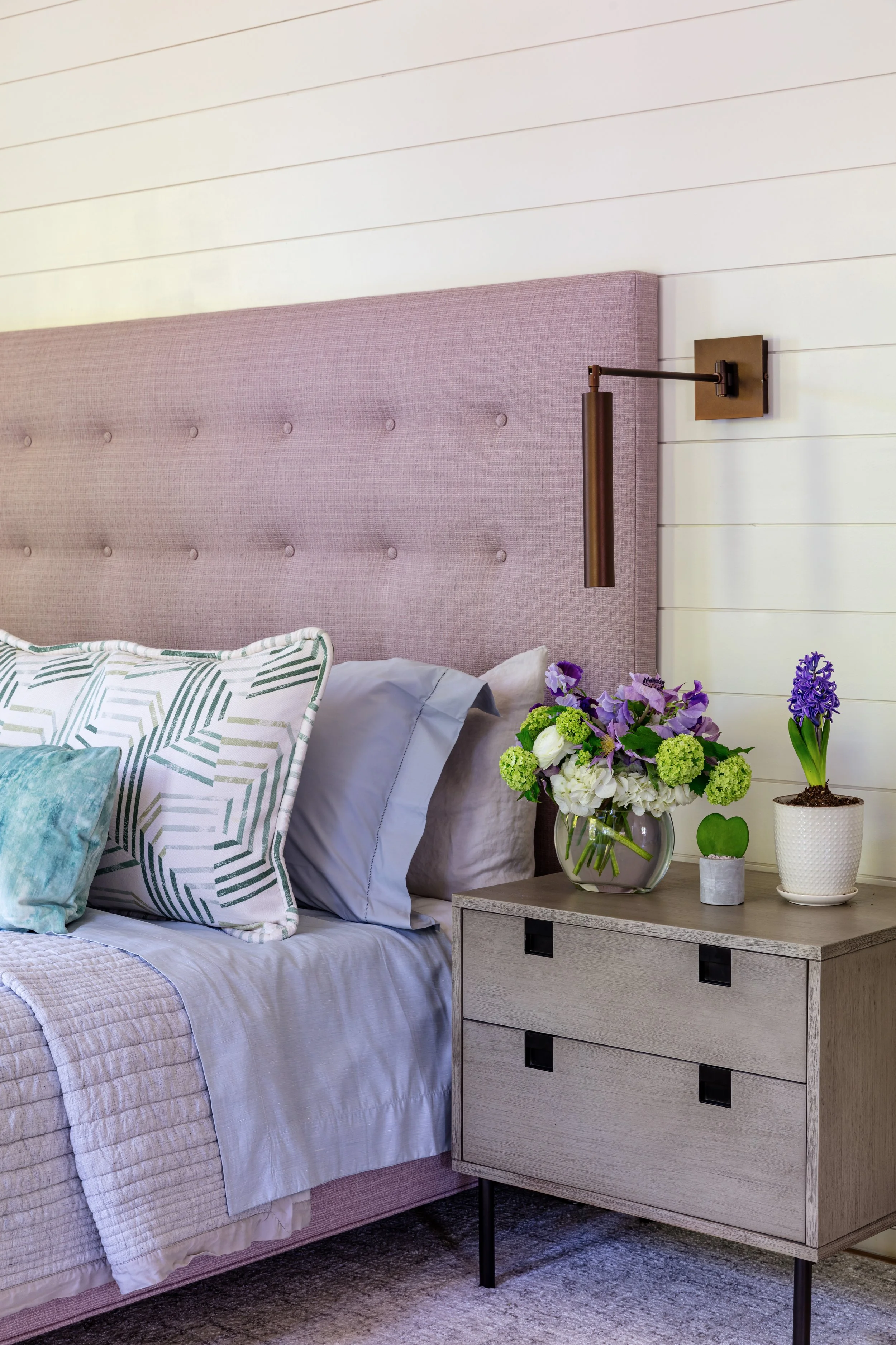 bedroom-detail-of-purple-tufted-headboard-gray-quilted-bedding-green-floral-drapery-rattan-chandelier-and-modern-art-decor-for-interior-design-inspiration.jpg