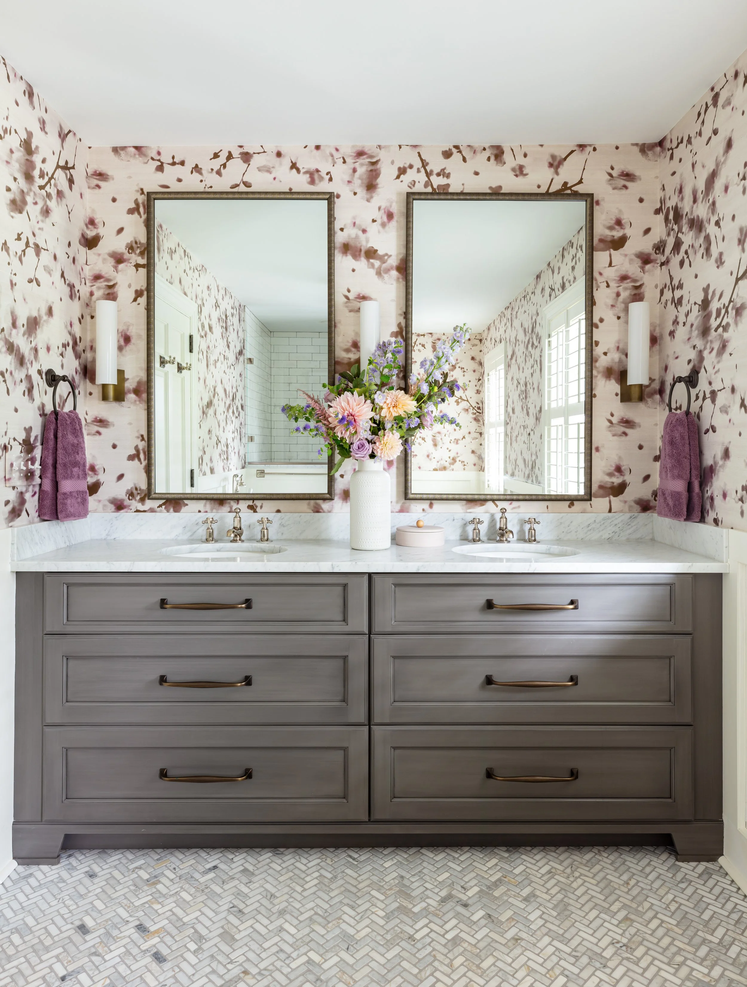 Elegant double vanity bathroom with gray drawers, marble countertop, floral wallpaper, and stylish sconces for luxurious daily routines