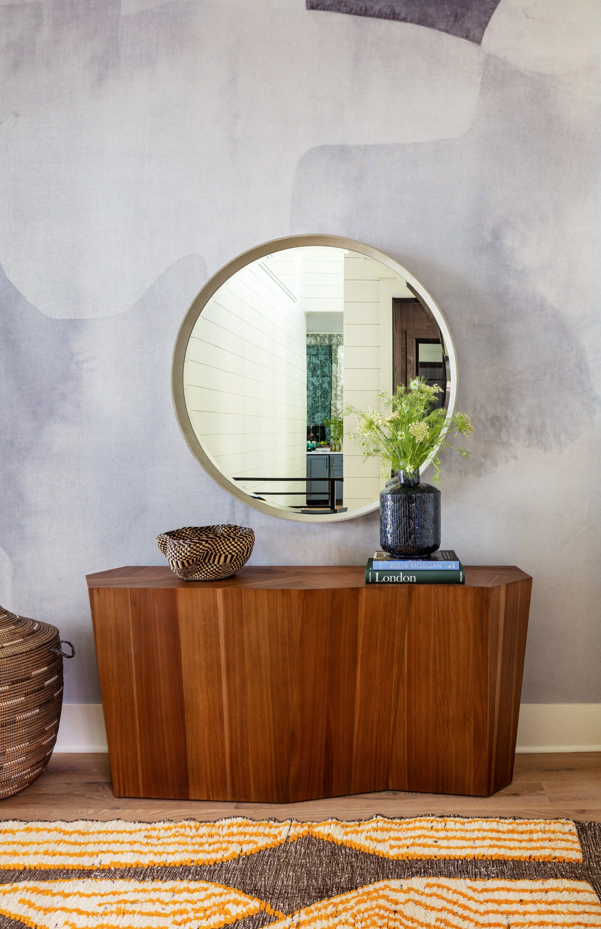 Mid-century modern entryway with warm wood console, round gold-framed mirror, woven basket, blue vase & books on patterned orange rug for stylish home décor inspiration