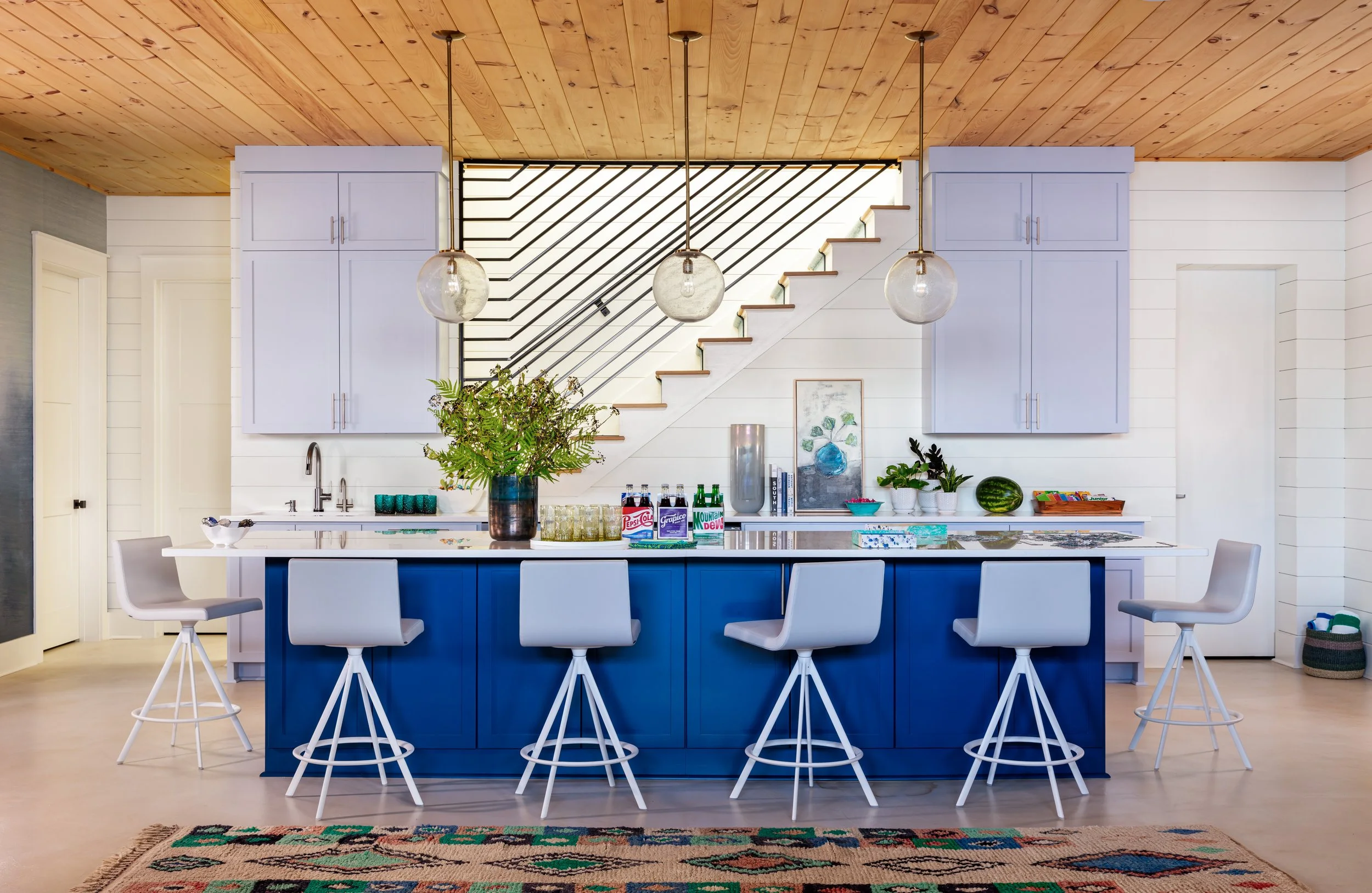 Modern kitchen with vibrant blue island, white cabinets, pendant lights, gray bar stools, and open staircase view for stylish interior design inspiration