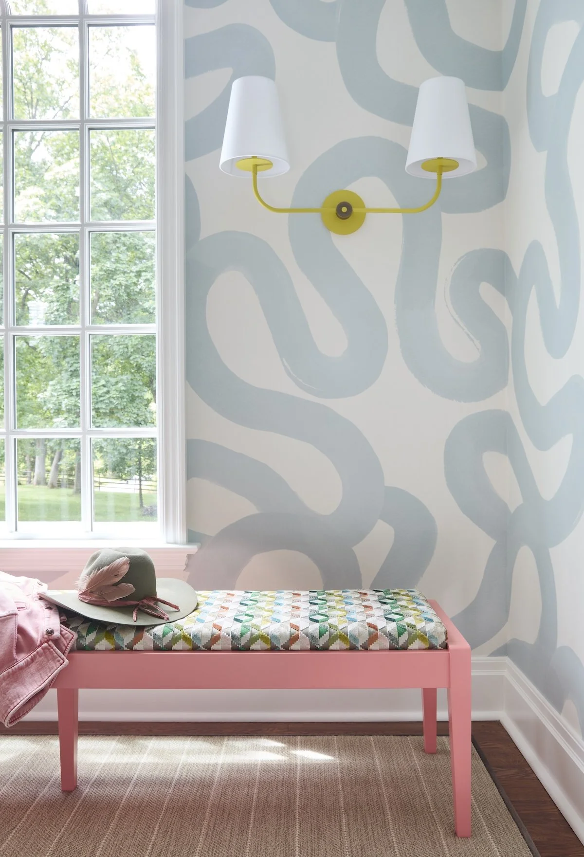 Pink upholstered bench with colorful geometric fabric under yellow double sconce and blue abstract wallpaper in bright window nook by Betsy Wentz