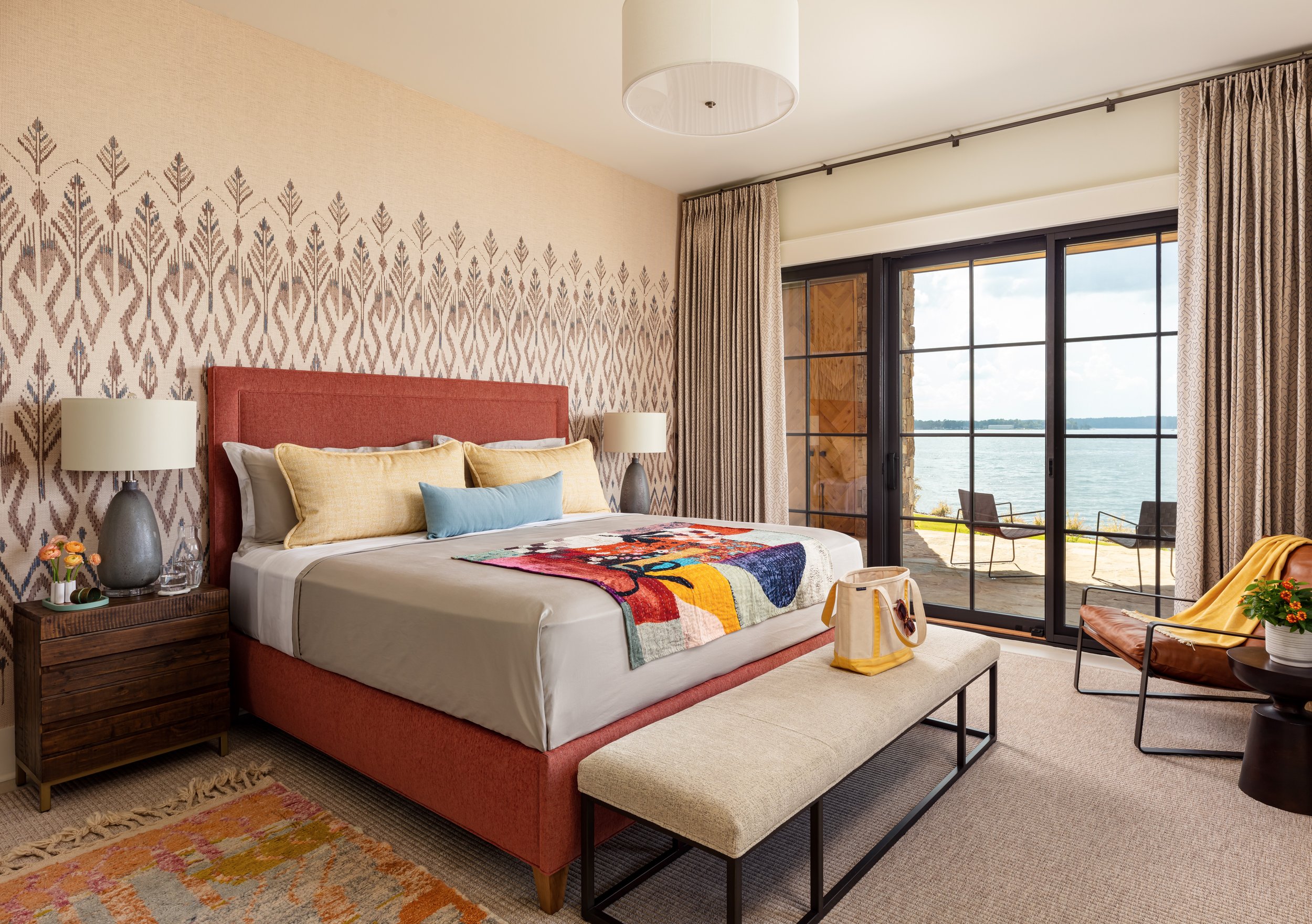 Stylish waterfront bedroom with upholstered bed, patterned wallpaper, modern lamps, and glass patio access for luxe interior design inspiration.
