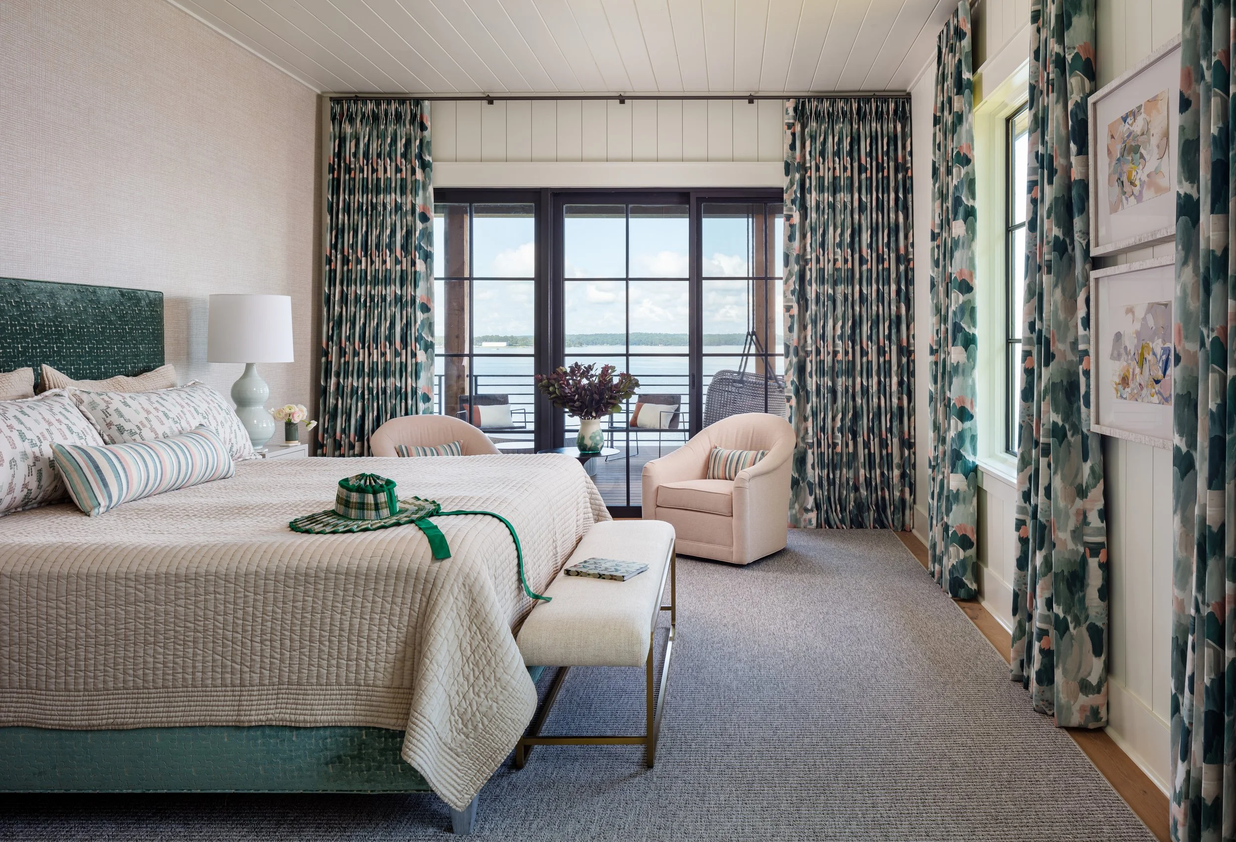Coastal-inspired bedroom with green upholstered headboard, patterned curtains, beige bedding, waterfront view, stylish armchair & decorative hat for chic interior design inspiration