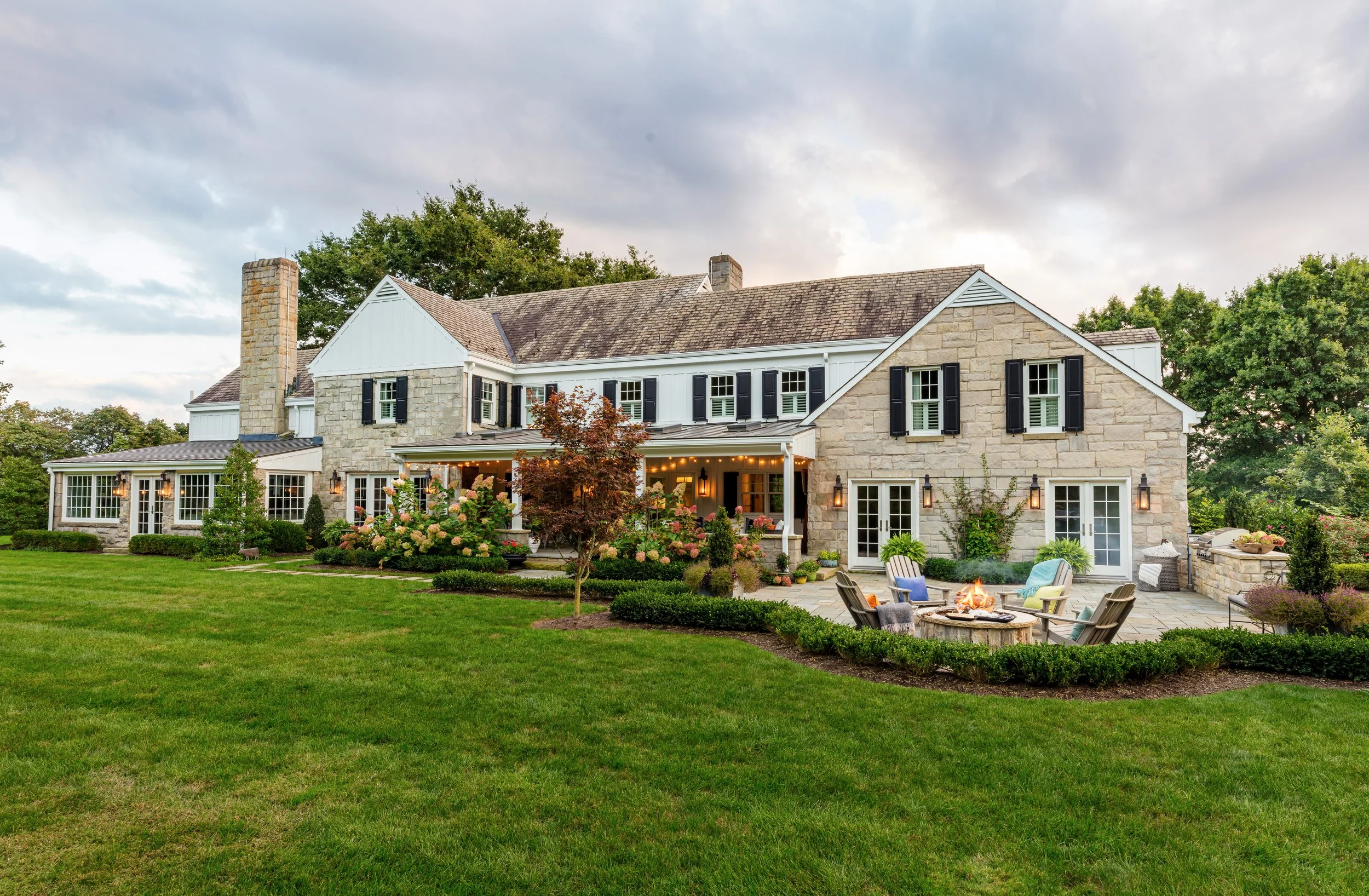 Elegant stone country home with charming porch, lush landscaping, and outdoor fire pit seating for cozy family gatherings and stylish entertaining