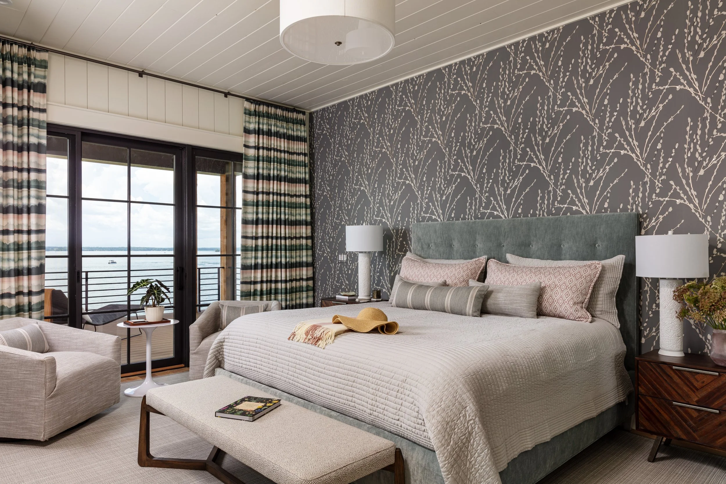 modern bedroom with patterned wallpaper, gray upholstered bed, cozy bedding, nightstand lamp, swivel chairs in front of large window with curtains