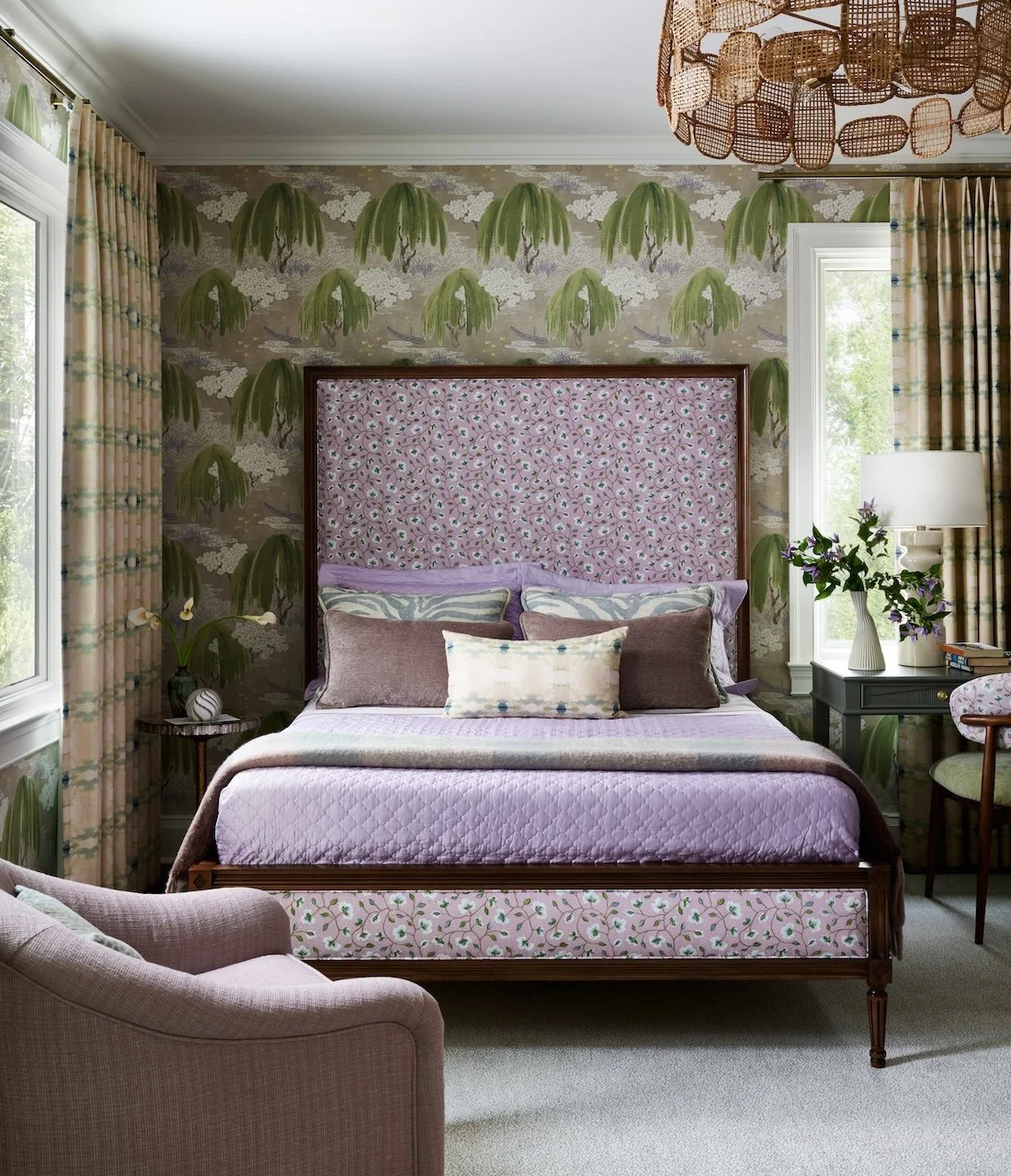 Lilac bedroom willow wallpaper floral upholstered bed patterned drapes rattan chandelier layered pillows mauve armchair
